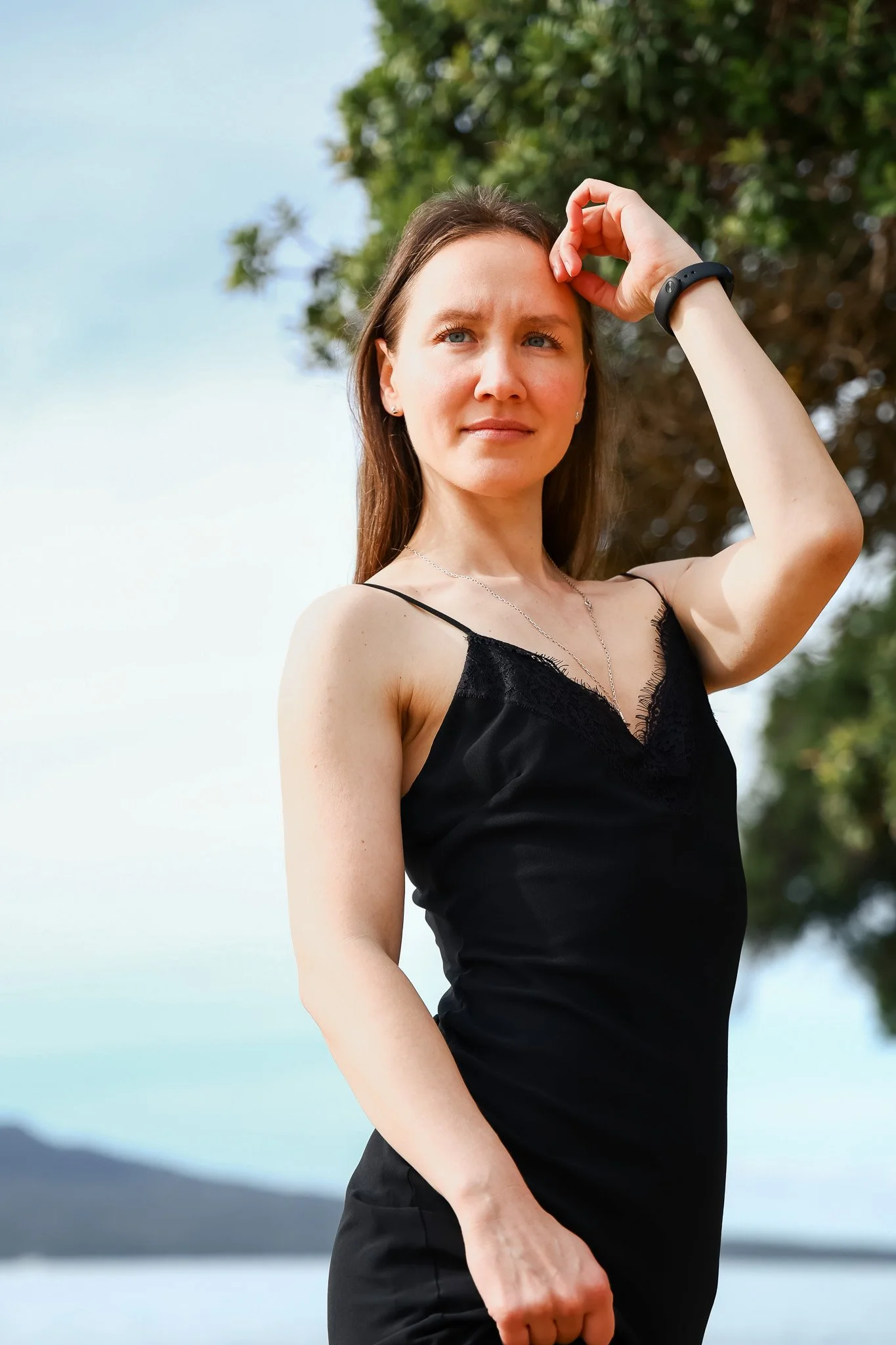 A woman with brown hair and blue eyes wearing a black top and a bracelet, standing outdoors near water and trees, looking into the distance with her hand touching her forehead.