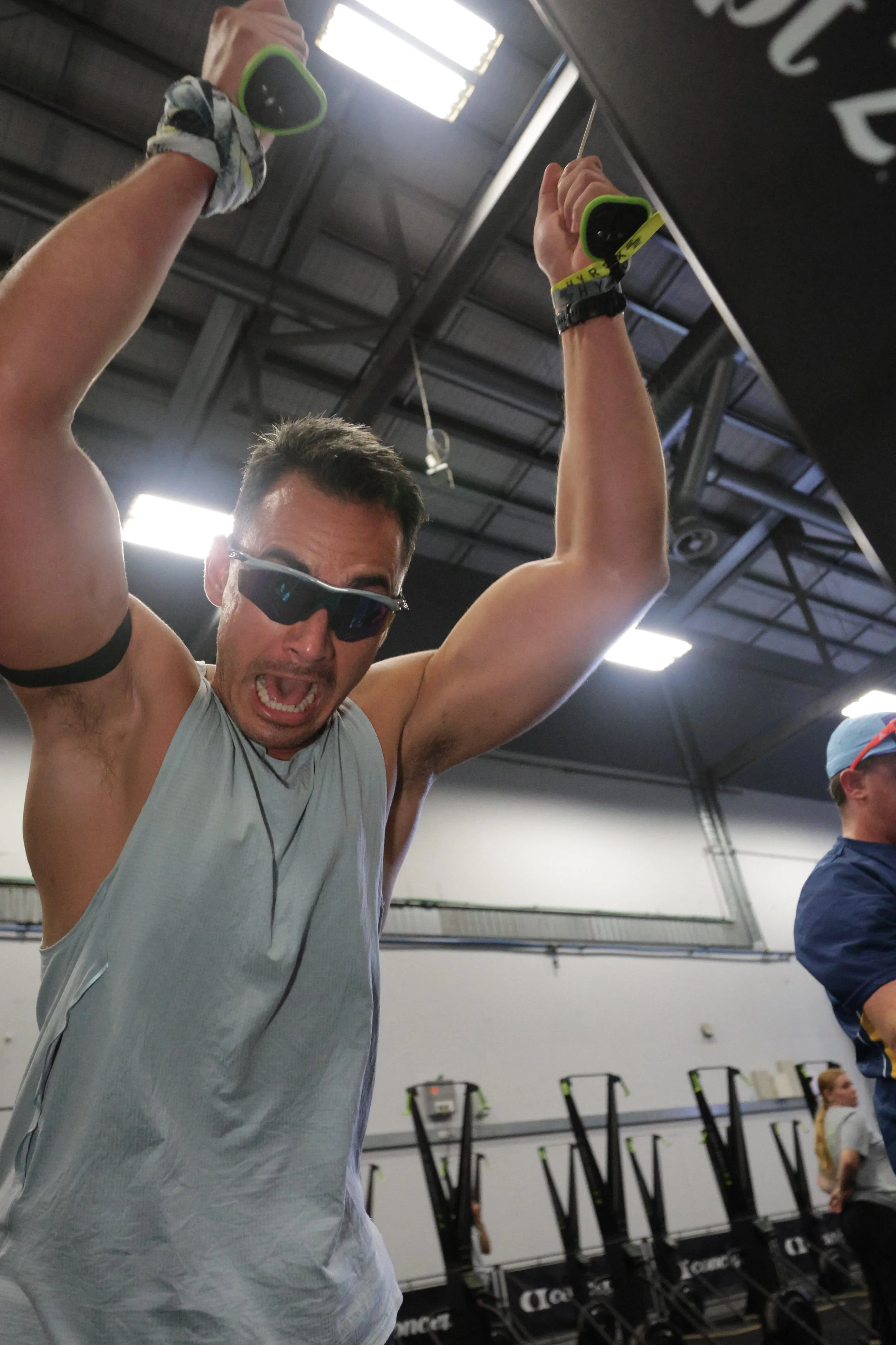 A man with sunglasses and a gray sleeveless shirt lifting weights in a gym, with a woman in the background, fitness equipment, and stacked cardio machines.