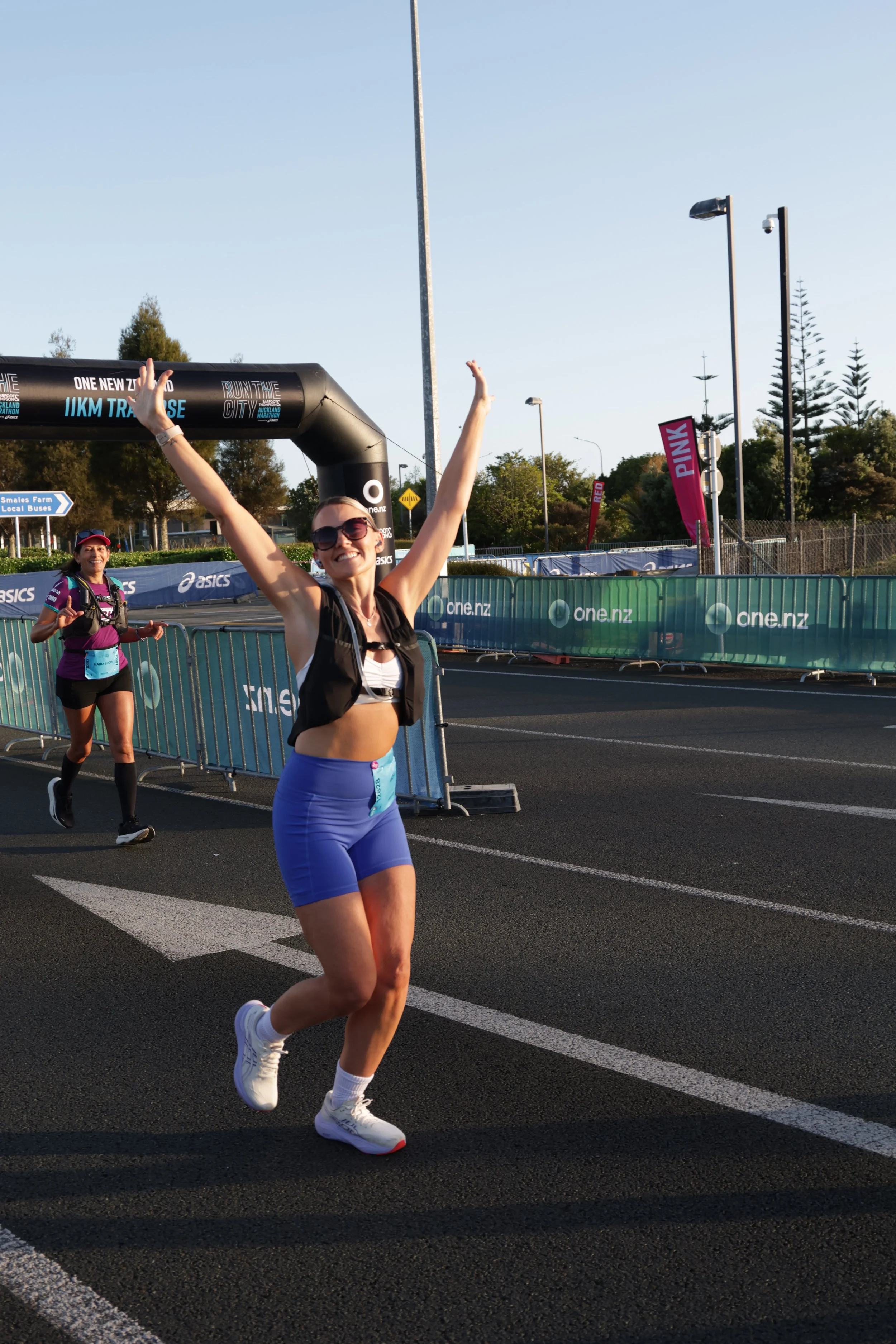 A woman runner in athletic attire, shorts and crop top, celebrating finishing a race with her arms raised, running on a marked road near a race finish line with people and banners in the background.
