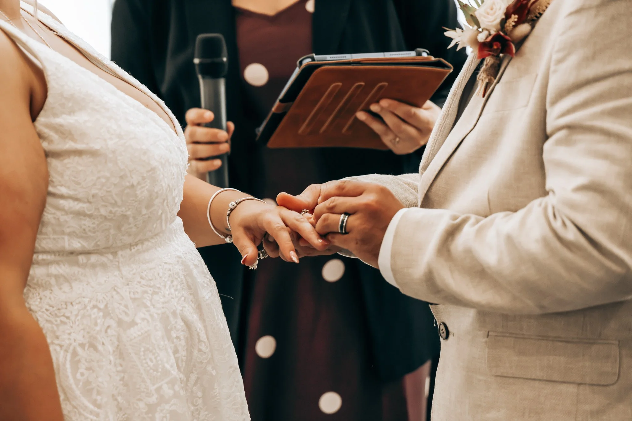 A couple holding hands during a wedding ceremony, with the bride wearing a white lace wedding dress and jewelry, and the groom wearing a cream-colored suit with a boutonniere, while an officiant reads from a folder.