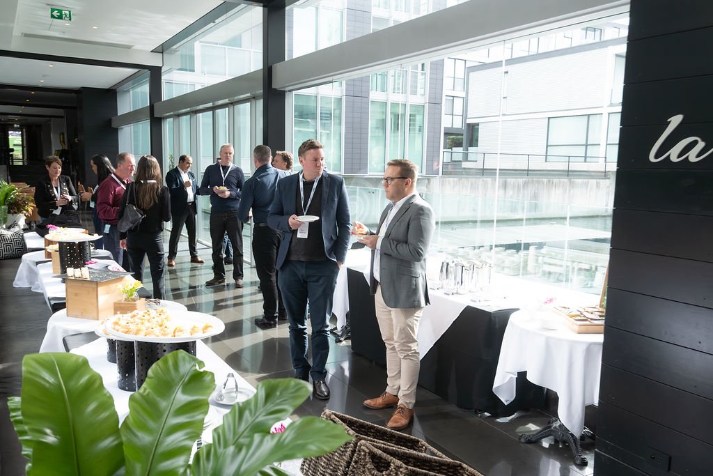 Business professionals at a networking event in a modern, glass-walled conference room with snack tables.