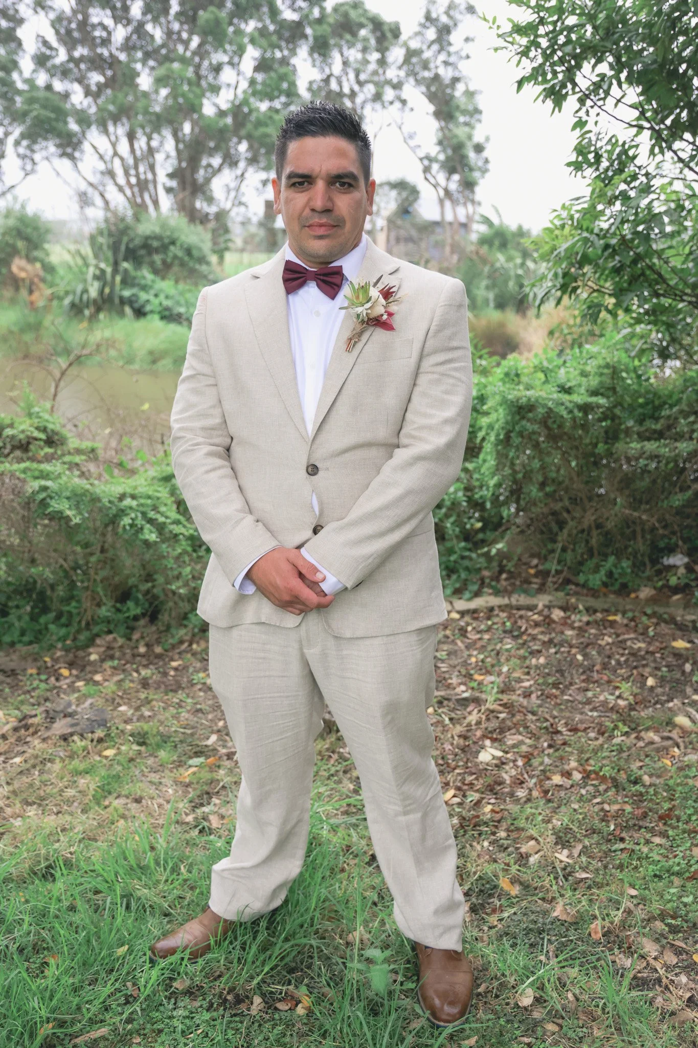 A man dressed in a beige suit with a matching tie and a boutonniere on his lapel stands outdoors on grass and dirt with greenery and trees in the background.