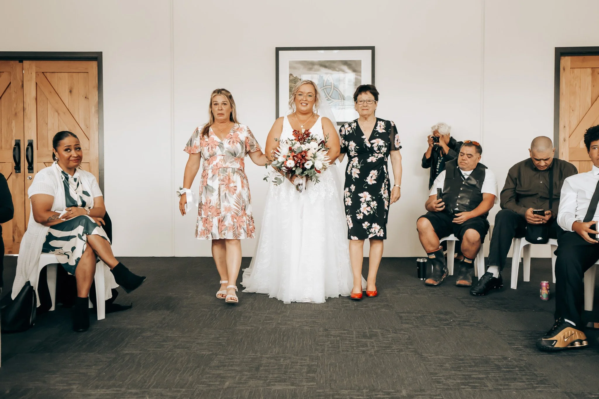 A bride in a wedding dress holding a bouquet, flanked by two women, at a wedding ceremony with seated guests.