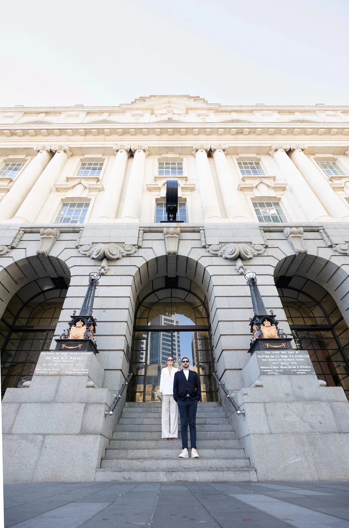 Two people in formal attire standing on a stone staircase in front of a grand, historic building with large columns and ornate details. The building has several stories and tall windows.
