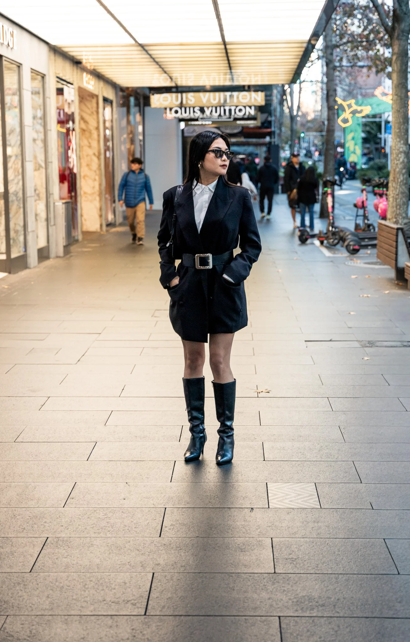 A woman wearing a black blazer dress, tall black boots, and sunglasses stands on a city sidewalk near a Louis Vuitton store with other pedestrians in the background.