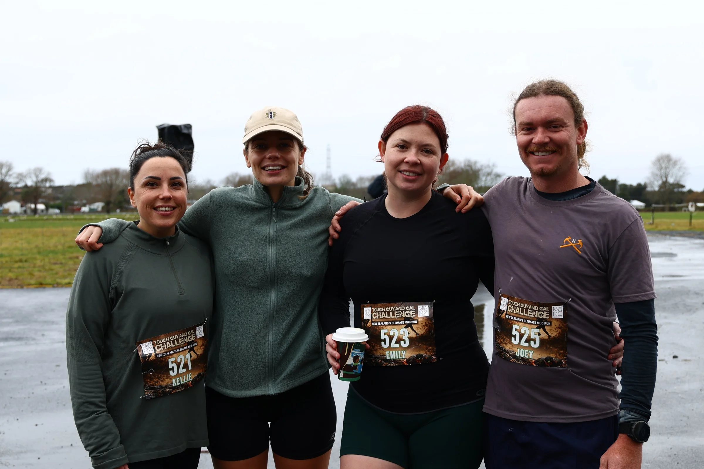 Four smiling people standing outdoors in a cloudy setting, each wearing a bib number, with two women and one man in the front and a woman in the back, embracing or having their arms around one another.