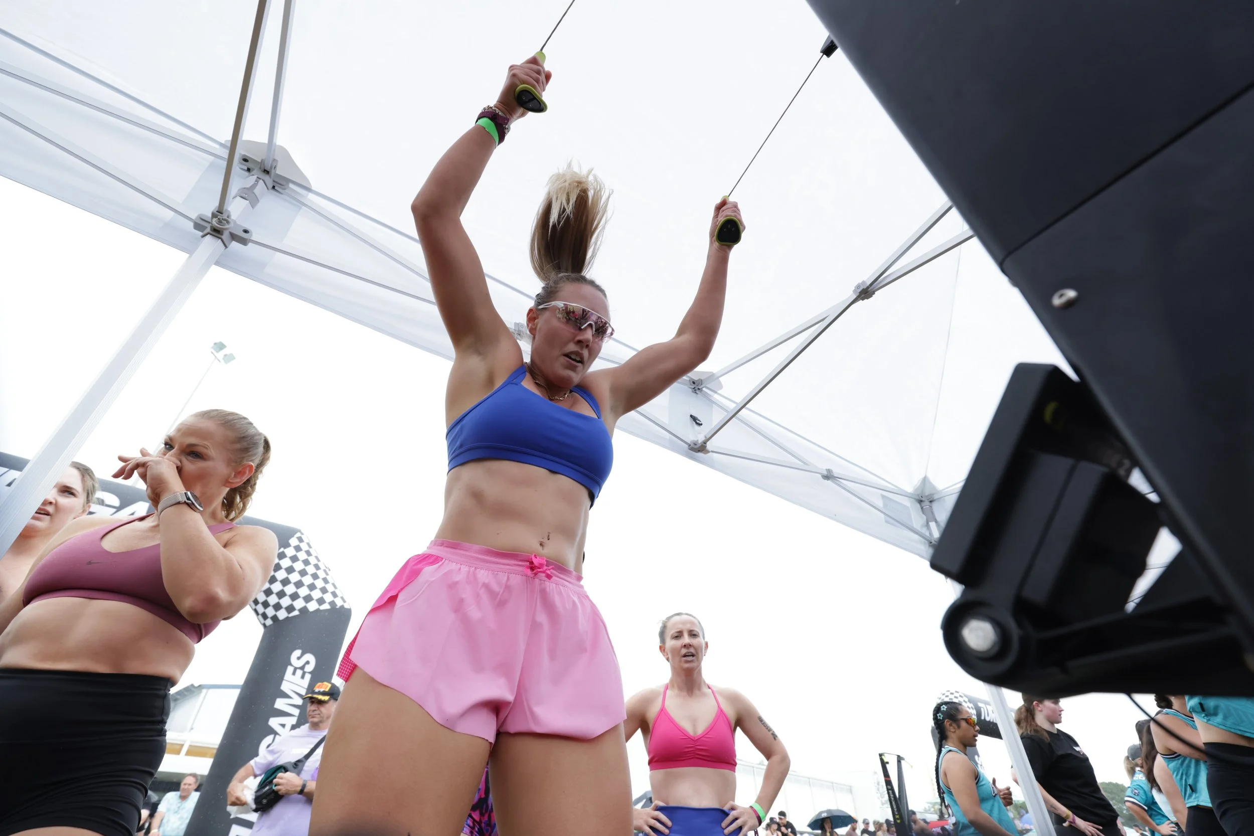 A woman in pink athletic shorts and a blue sports bra raising her arms above her head during a race or competition at an outdoor event. Several other women are standing around her, some wearing athletic gear, at a venue with tents and event banners.