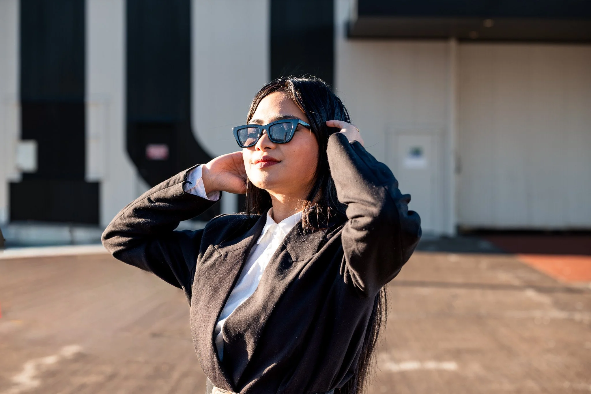 A woman with black hair wearing sunglasses, a black blazer, and a white shirt poses outdoors with her hands near her head. The background shows a building and a parking lot.