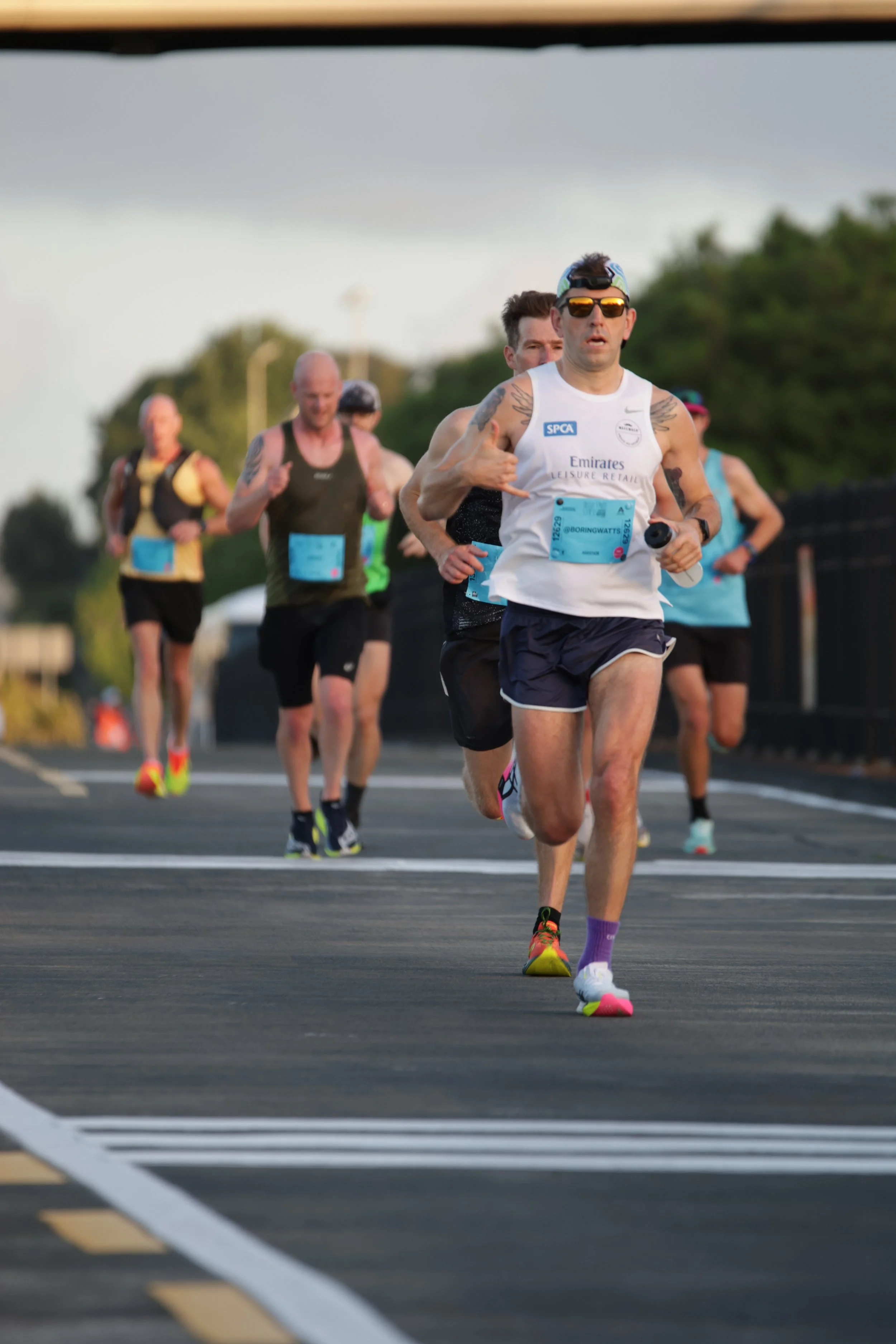A group of runners participating in a race, with one man in the foreground wearing sunglasses, a baseball cap, and a white tank top, making a hand gesture. Other runners are visible behind him on an outdoor road.
