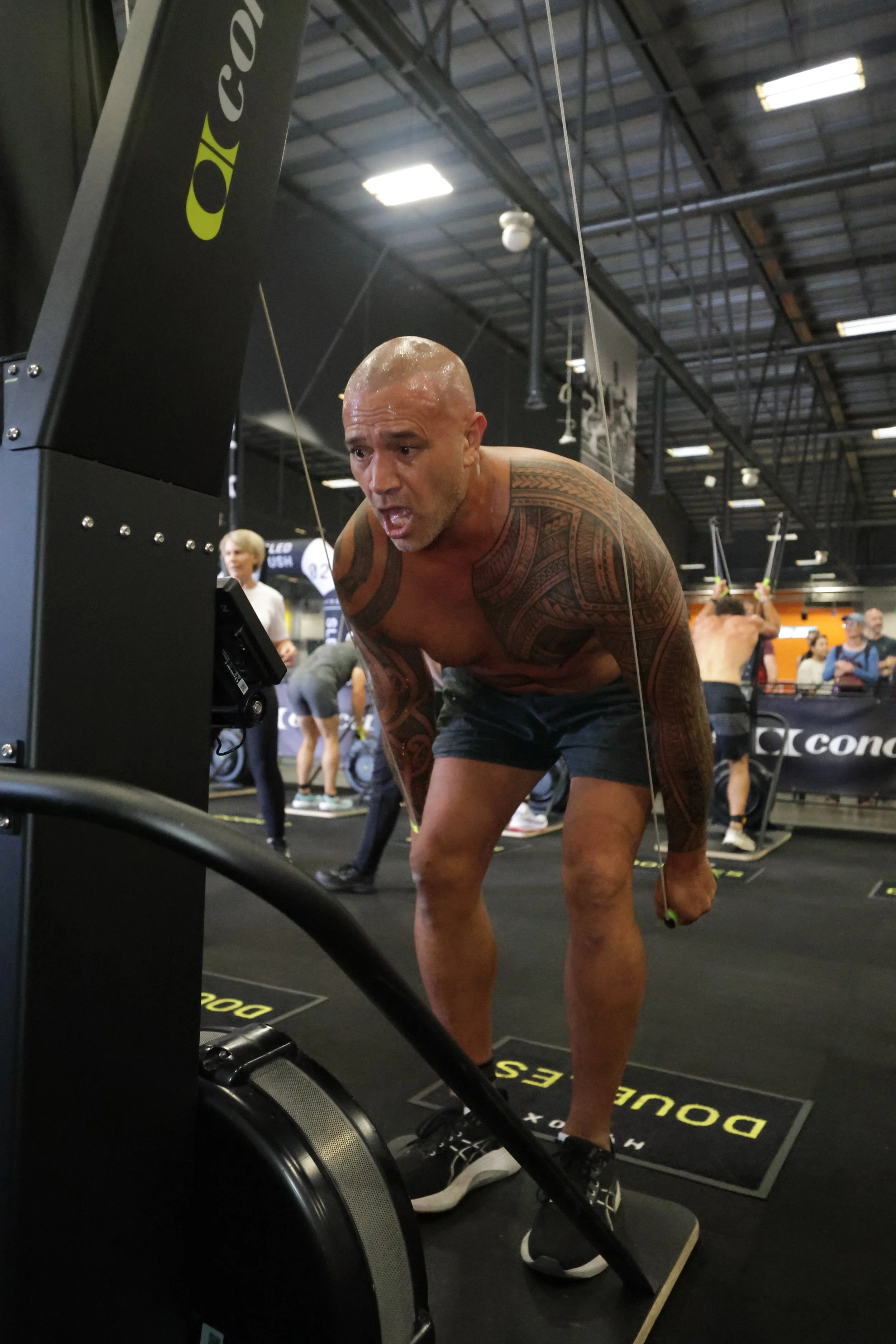 A shirtless man with tattoos bent over, holding a jump rope, in a gym with other people exercising in the background.