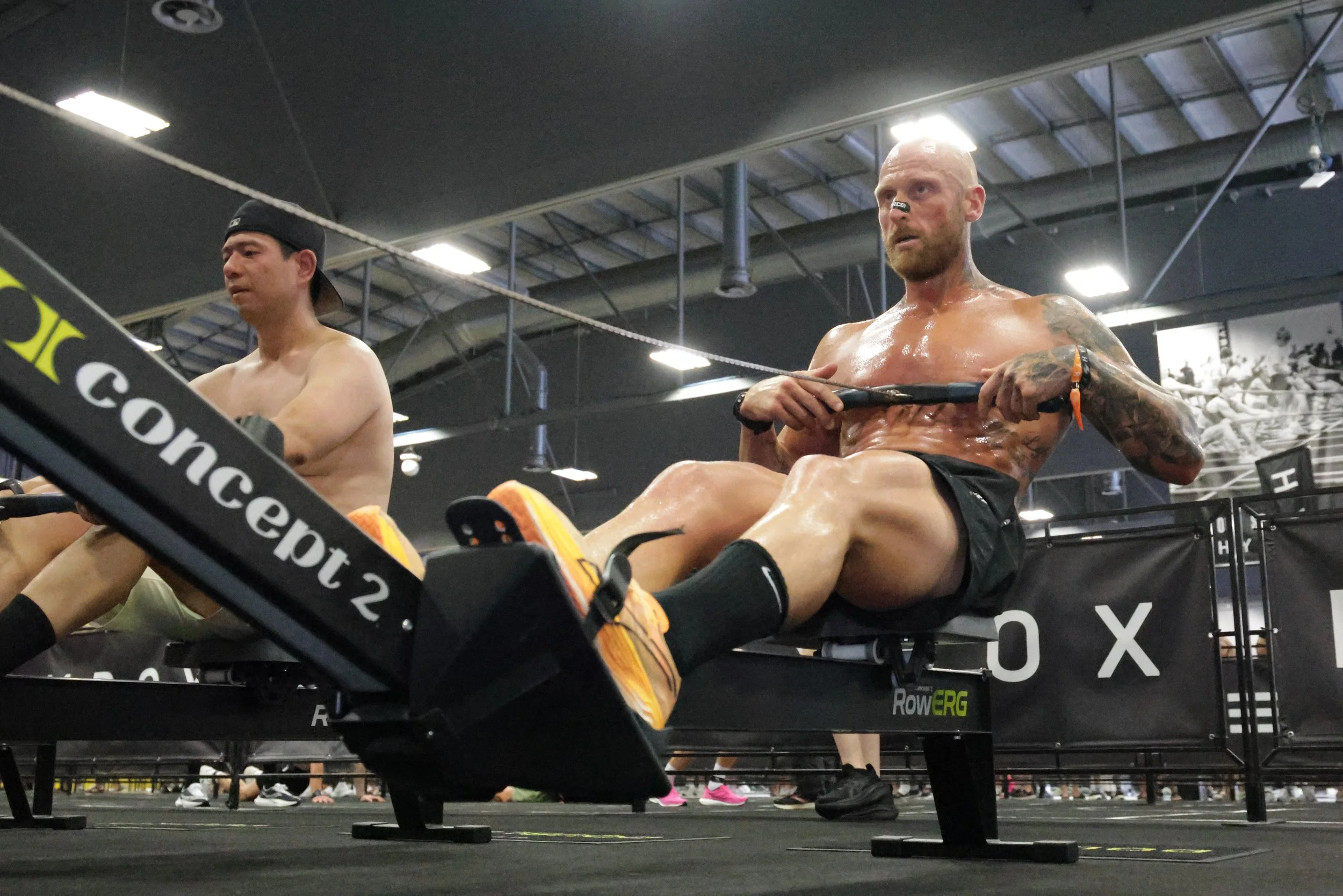 Two shirtless men are participating in a rowing exercise inside a gym. One is wearing a black cap and the other has a tattooed arm, and both are pulling on rowing machines.