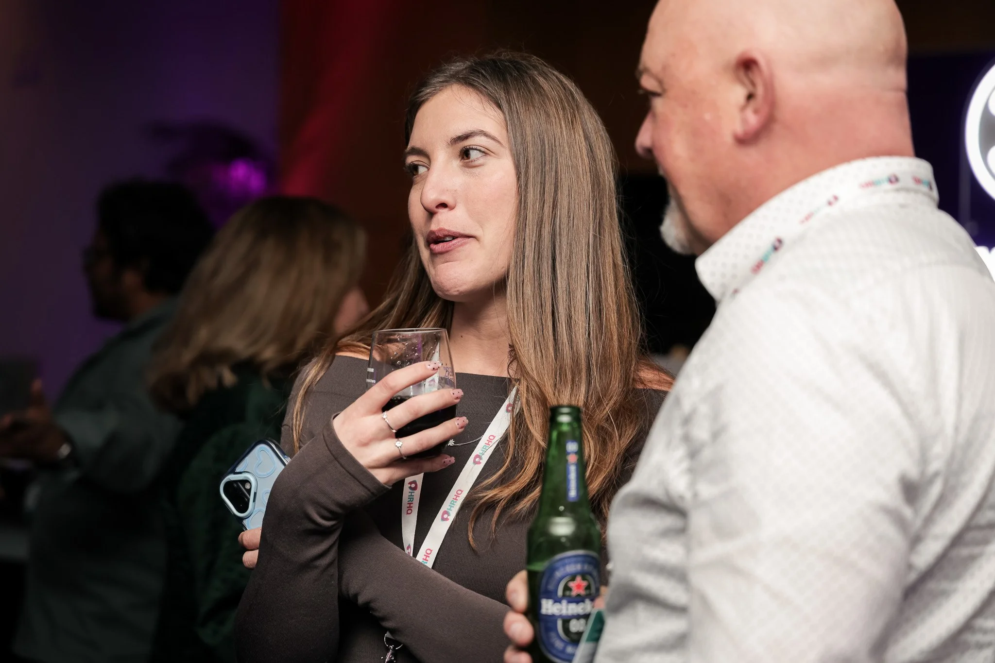 A woman with long brown hair holding a glass of dark beverage and talking to a man with a bald head holding a Heineken beer bottle at a social event.