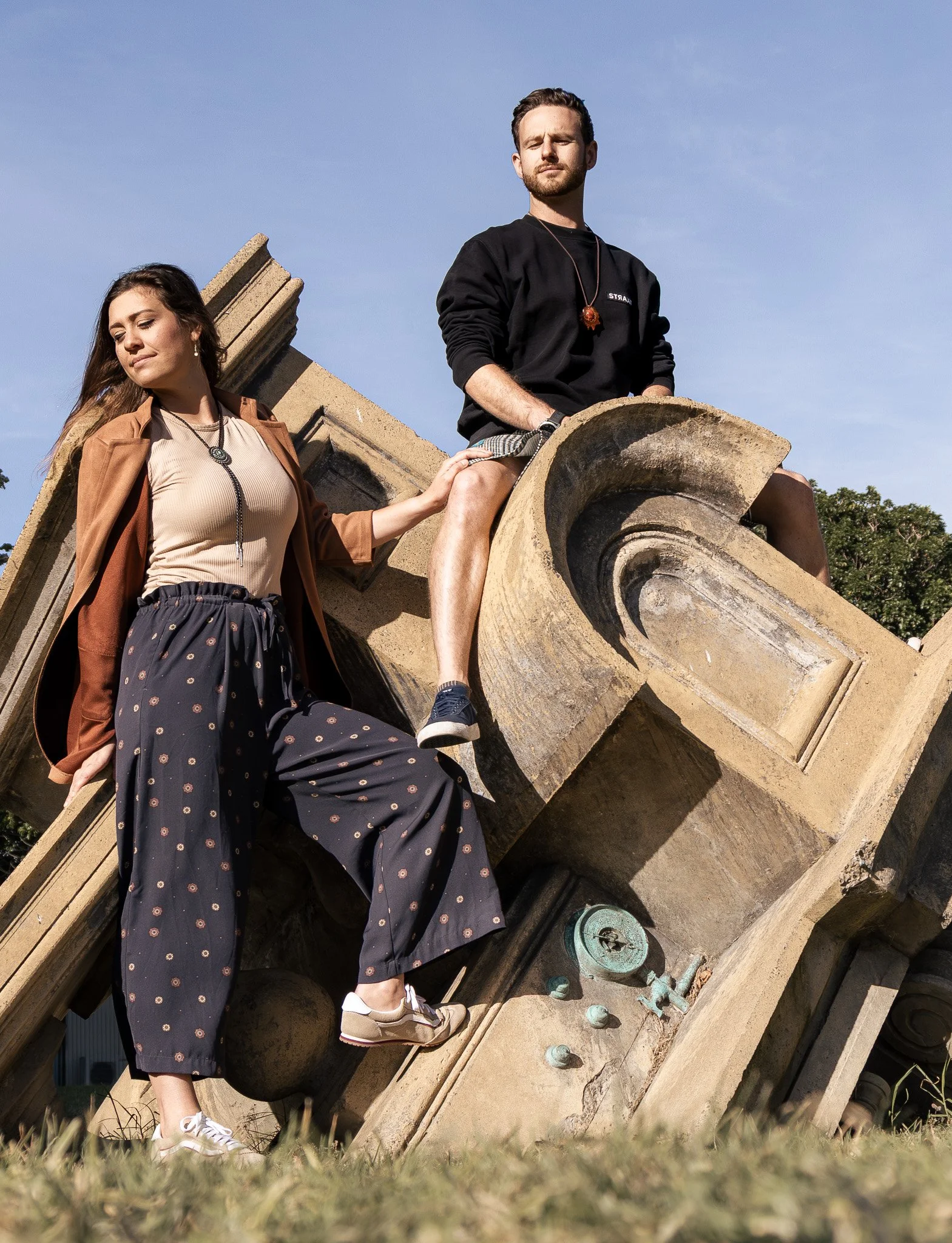 A young man and woman sit on a stone sculpture resembling an ear and a head, outdoors with blue sky and trees in the background. The man wears a black sweatshirt, shorts, and sneakers, while the woman wears a beige top, patterned pants, and a brown j