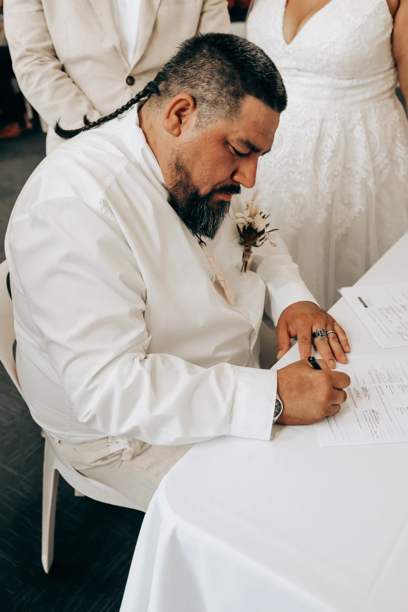A man with dark hair and a beard, dressed in white, is signing a document at a white round table during a wedding ceremony. Behind him, partially visible, are a woman in a white wedding dress and another man in a light-colored jacket.