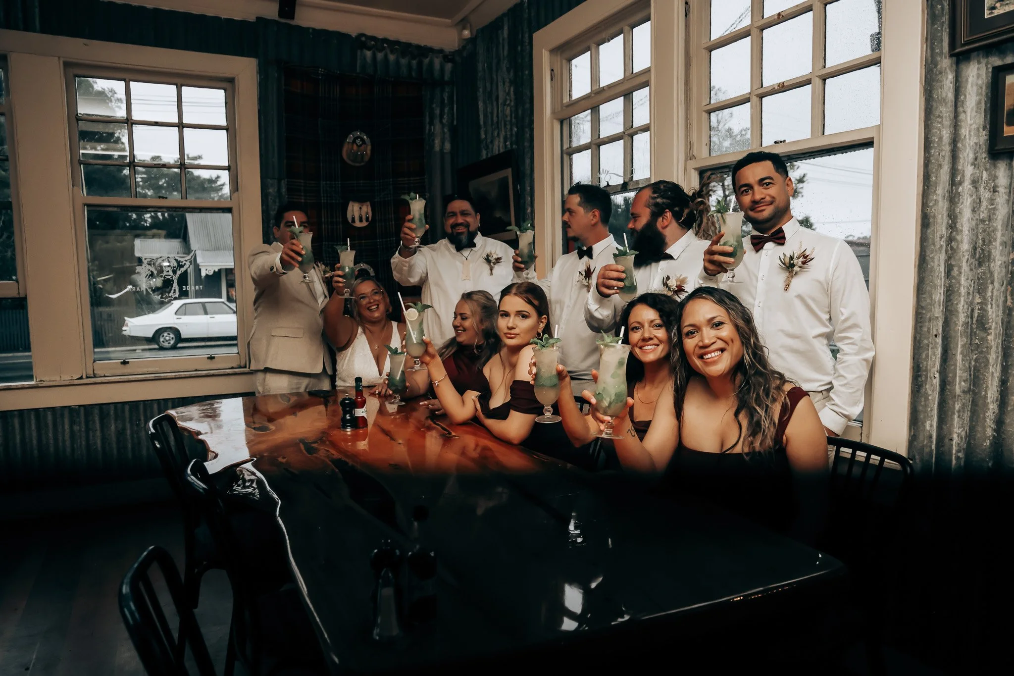 Group of people celebrating at a party, holding up drinks and smiling, in a room with large windows and a wooden table.