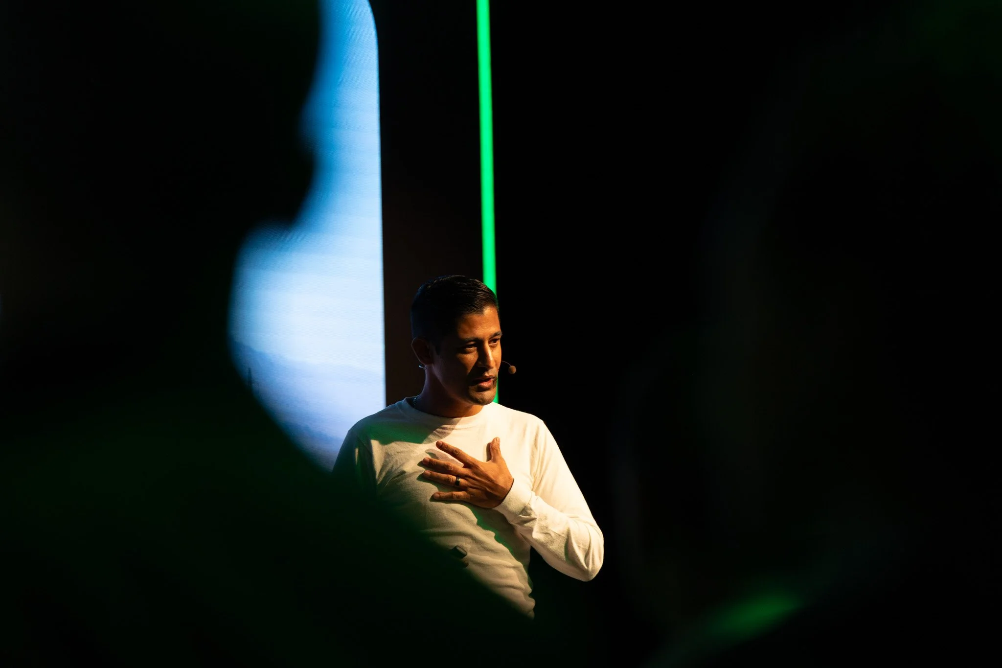A man wearing a white shirt with short hair, speaking on stage with a microphone, with one hand over his chest in front of a dark background, illuminated by green and blue lights.