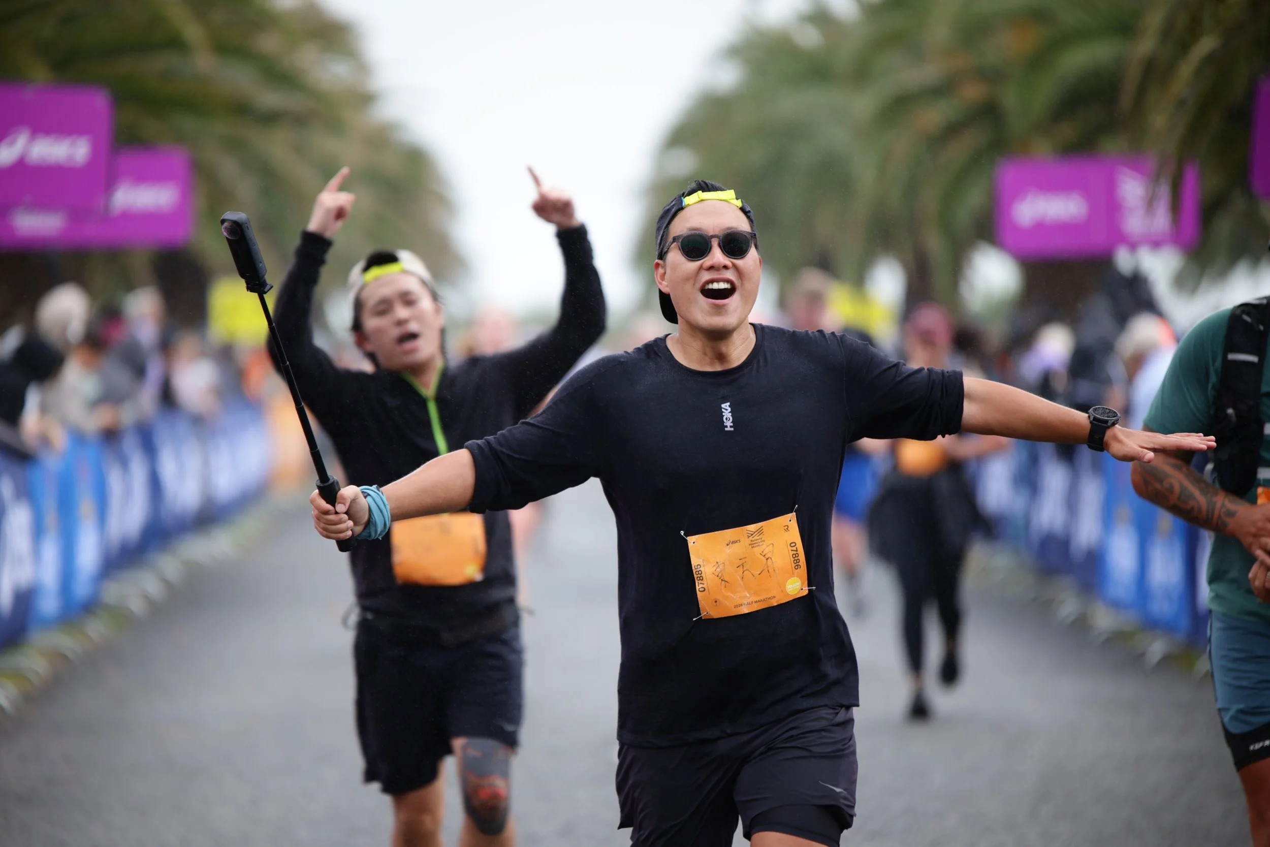 A man with sunglasses and a cap cheering with outstretched arms during a marathon, with other runners and spectators in the background.