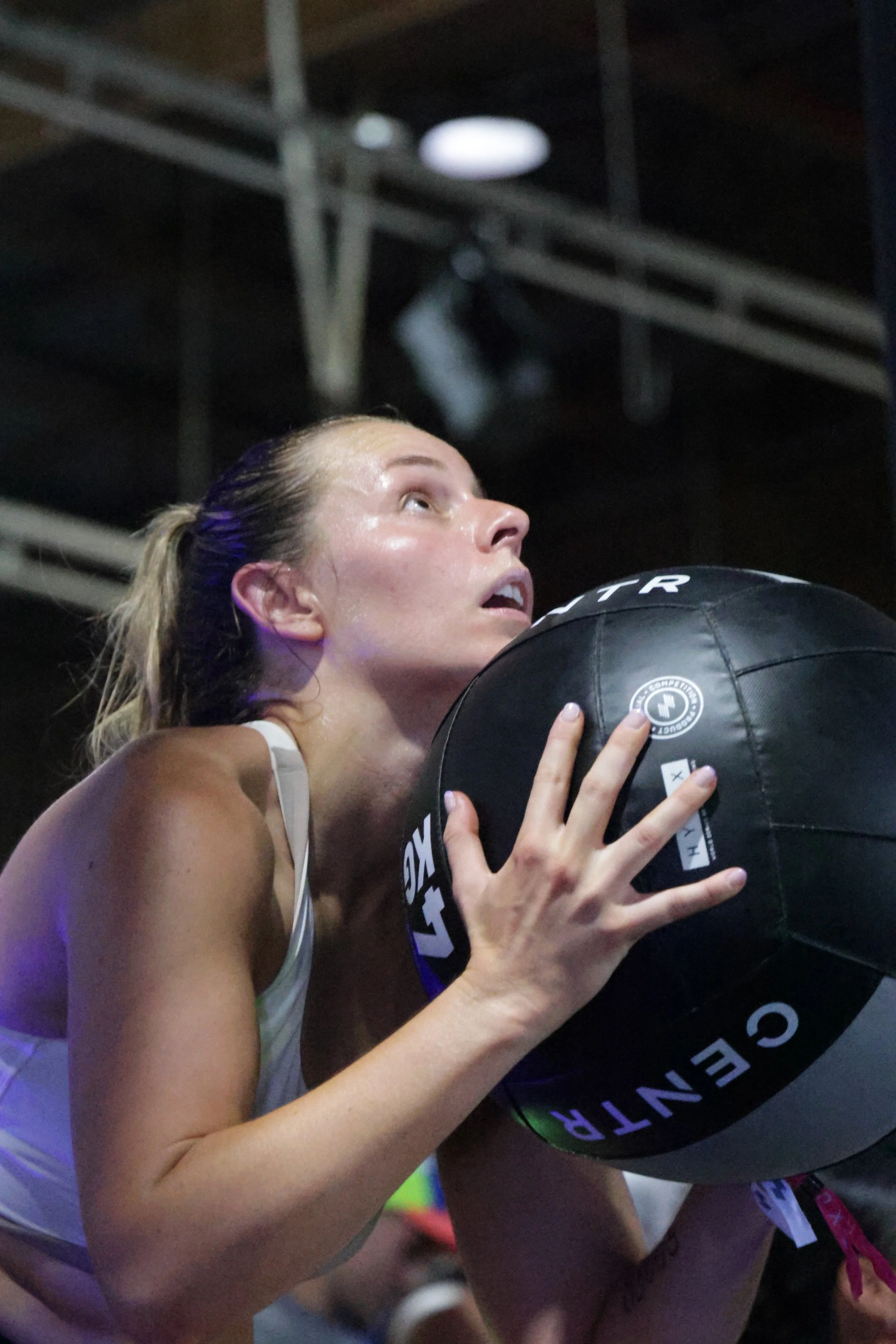A woman holding a black medicine ball close to her face during a workout, with her hair tied back and sweat on her face.