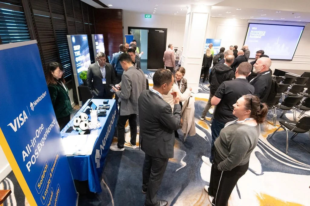People networking at a conference, with a booth displaying VISA and NewCharger branding, soccer balls on the table, and a large screen showing a presentation in the background.