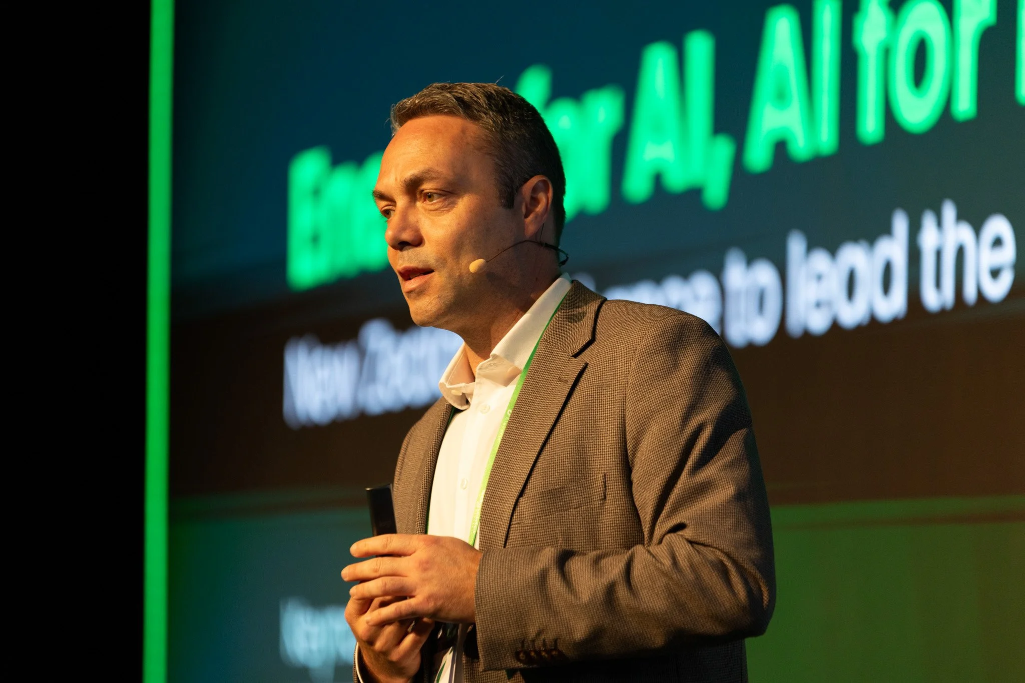 A man in a brown blazer and white shirt speaking into a microphone on stage, with a dark background and a large screen displaying green and white text behind him.