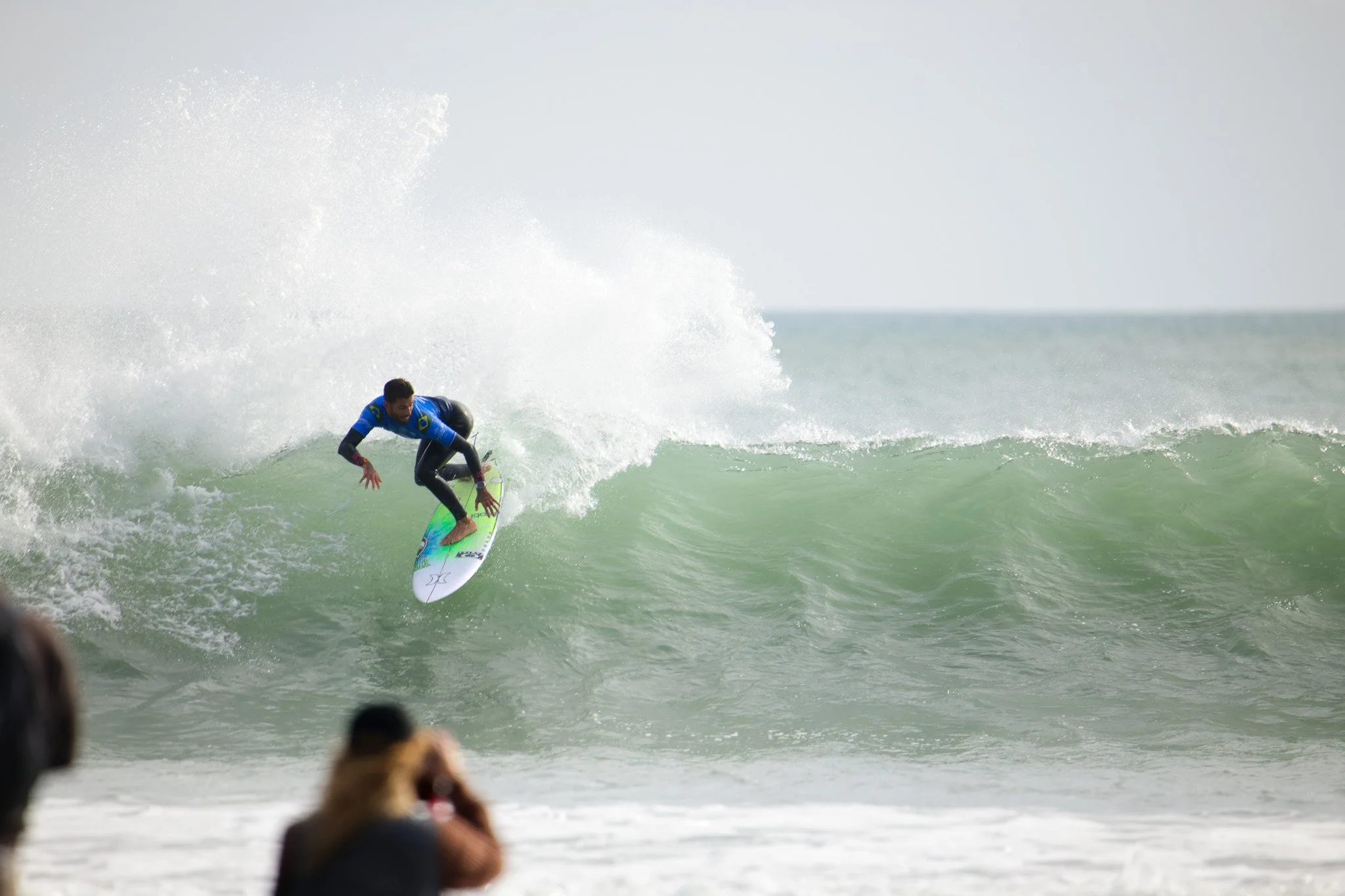 A male surfer riding a wave on a surfboard in the ocean, with a few spectators watching from the shore.