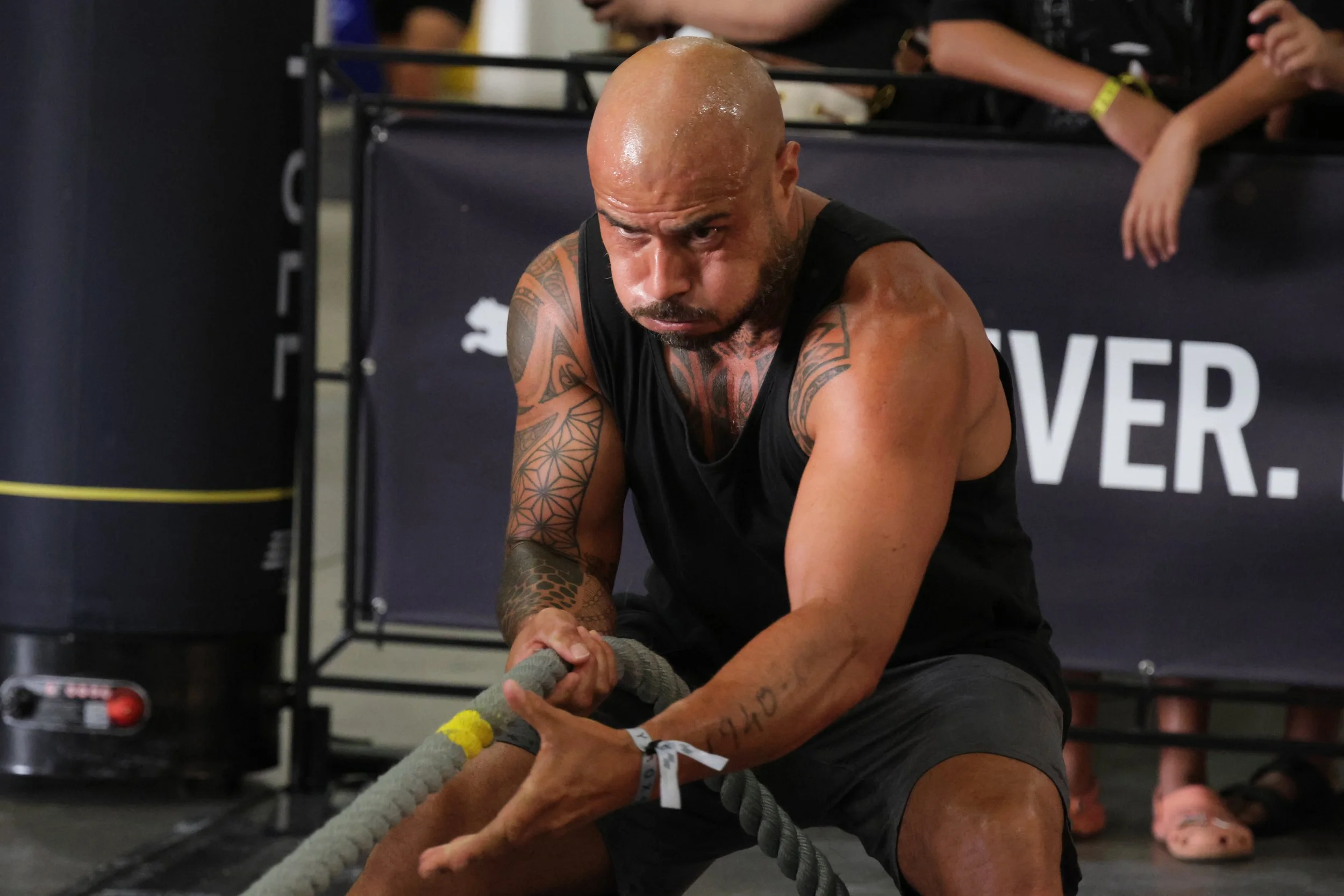 A muscular, tattooed man with a shaved head in black athletic clothing sitting on the floor, pulling a thick workout rope during a fitness event with onlookers in the background.