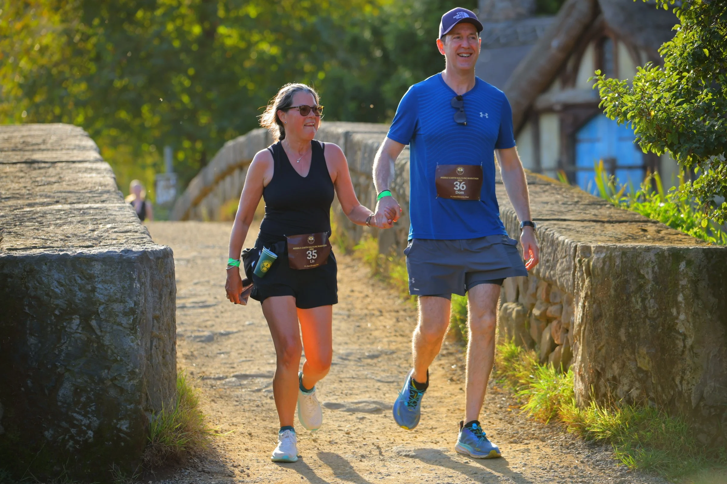Two people, a woman and a man, are running outdoors on a sunny day along a stone path with a stone wall on one side. The woman is wearing sunglasses, a black sleeveless top, and black shorts, while the man is wearing a blue shirt, blue shorts, and a 