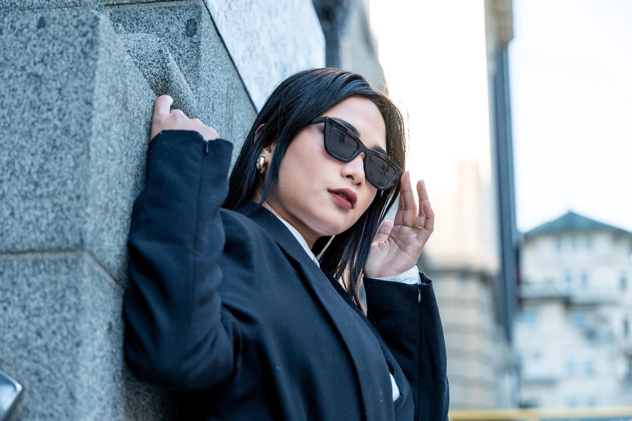 A woman with long dark hair, wearing dark sunglasses, a black blazer, and gold earrings, is sitting and leaning against a stone wall outdoors during daytime. She appears to be adjusting her sunglasses with her left hand.