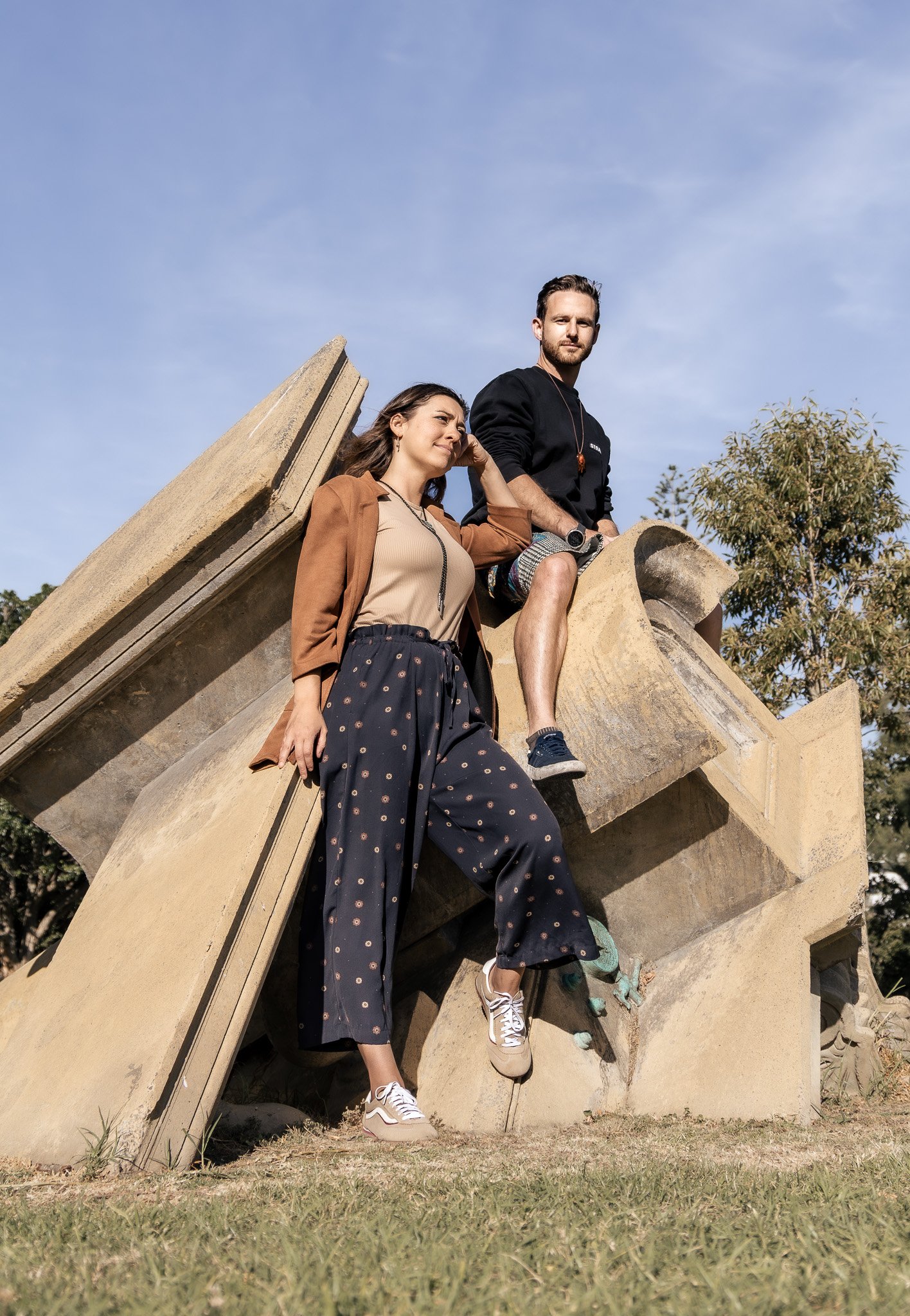 Two young adults sitting on a large concrete structure outdoors with a blue sky and trees in the background. The woman is leaning against the structure, while the man is sitting with one leg resting on it. They are dressed in casual clothing and snea
