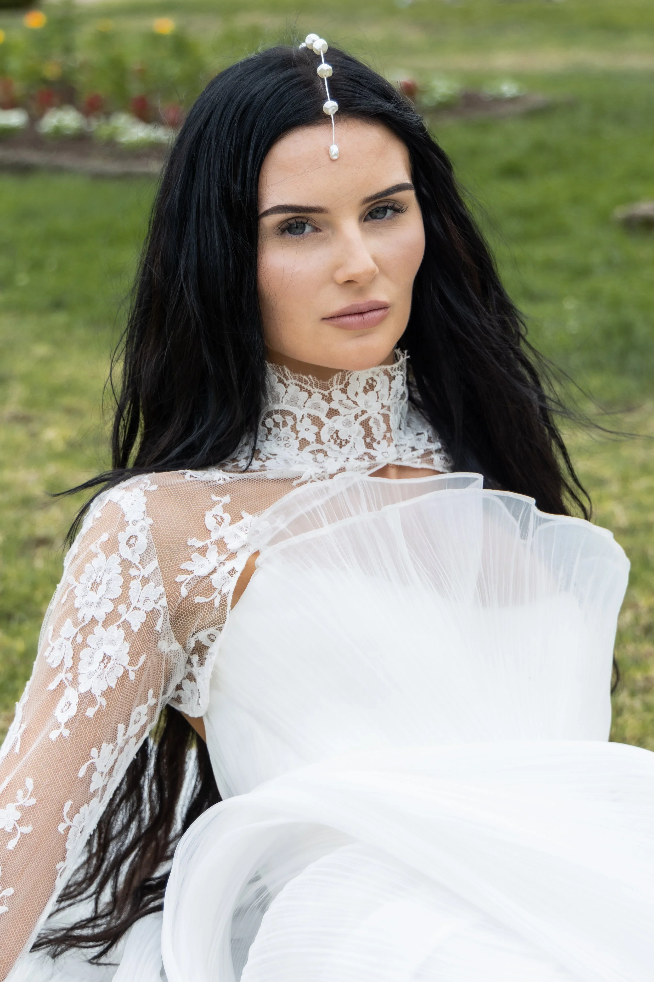 A woman with long black hair and blue eyes wearing a lace dress and holding a white floral fan outdoors.
