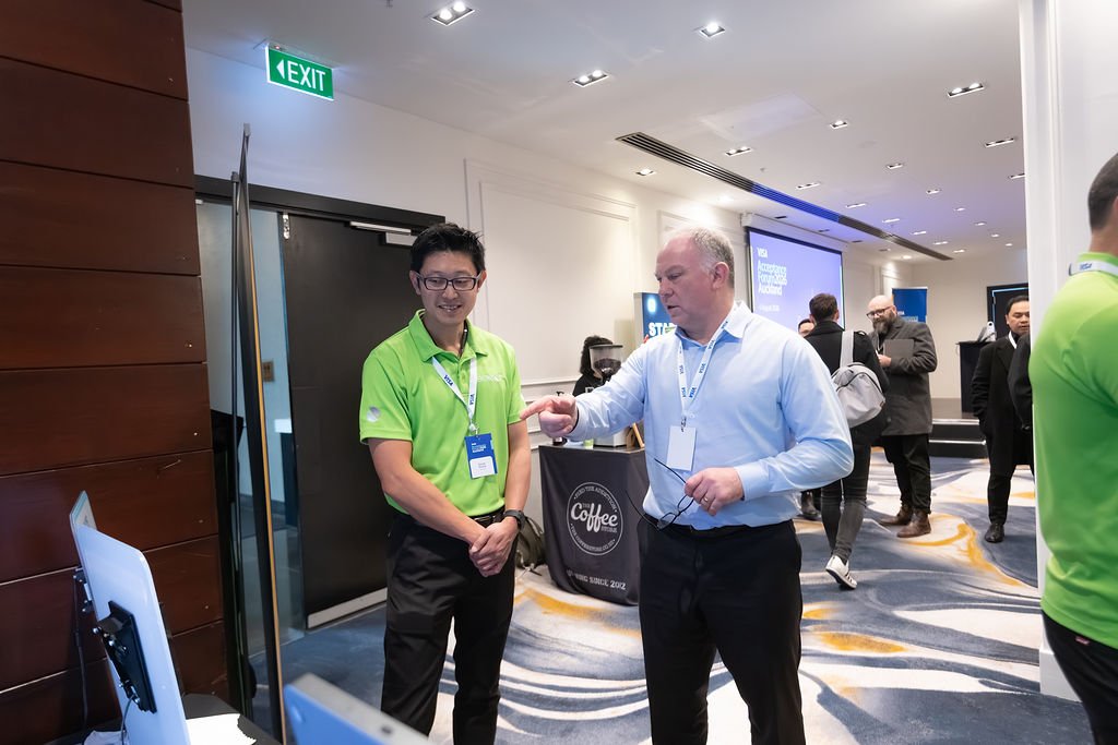 Two men are having a conversation at a conference. One man is wearing a bright green polo shirt and the other is wearing a light blue dress shirt. The room is filled with other attendees and displays in the background.