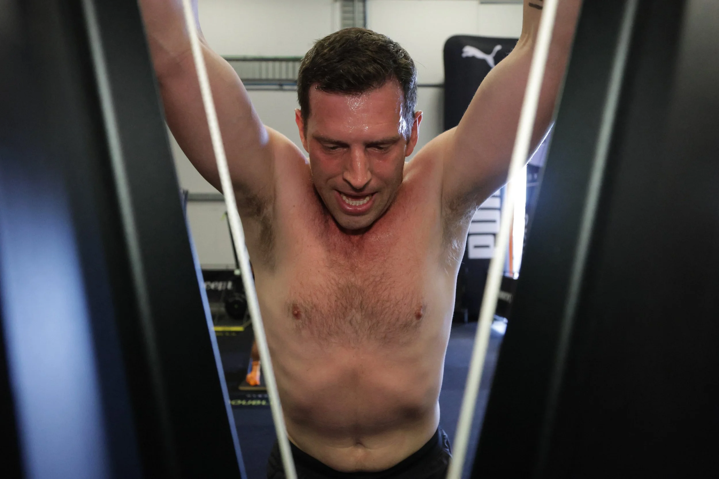 A shirtless man with short brown hair is performing a workout with his arms raised and bent at the elbows, seen through the bars of gym equipment. He appears to be sweating and focused while exercising in a gym setting.