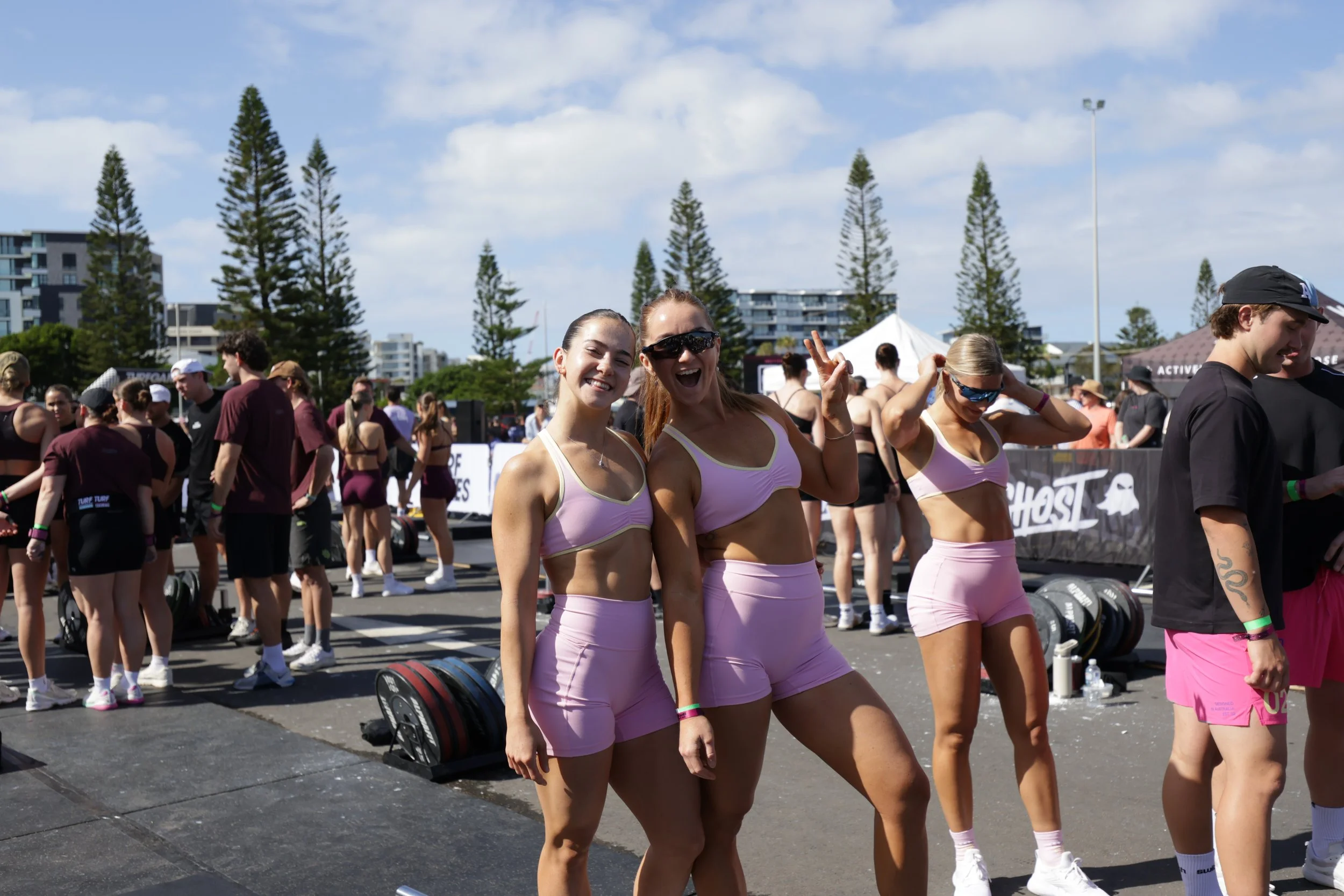 Smiling women in pink workout outfits at an outdoor fitness event, with a crowd in the background and tall trees against a partly cloudy sky.