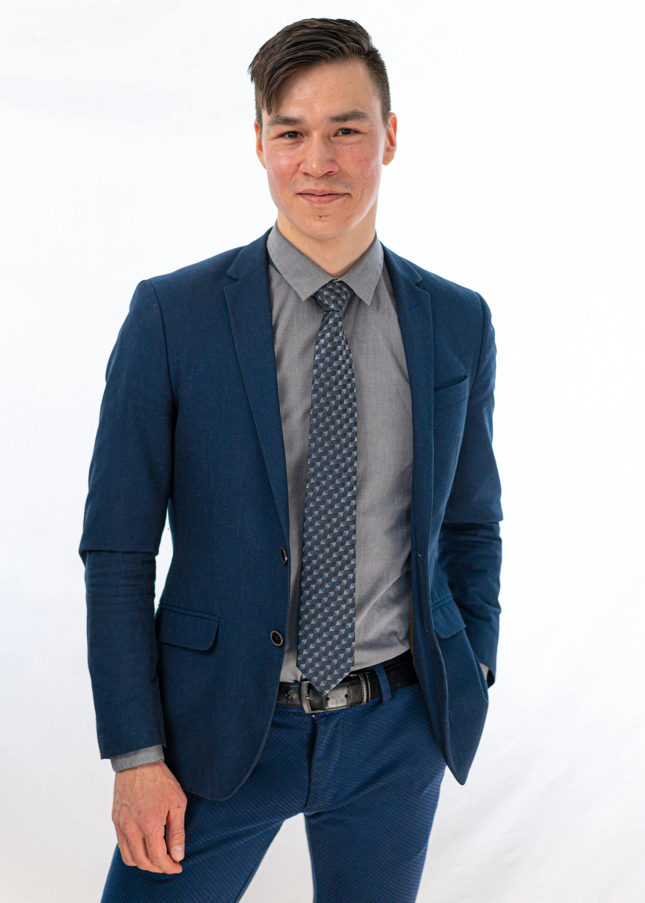 A man in a dark blue suit, gray shirt, and patterned tie standing against a plain white background.
