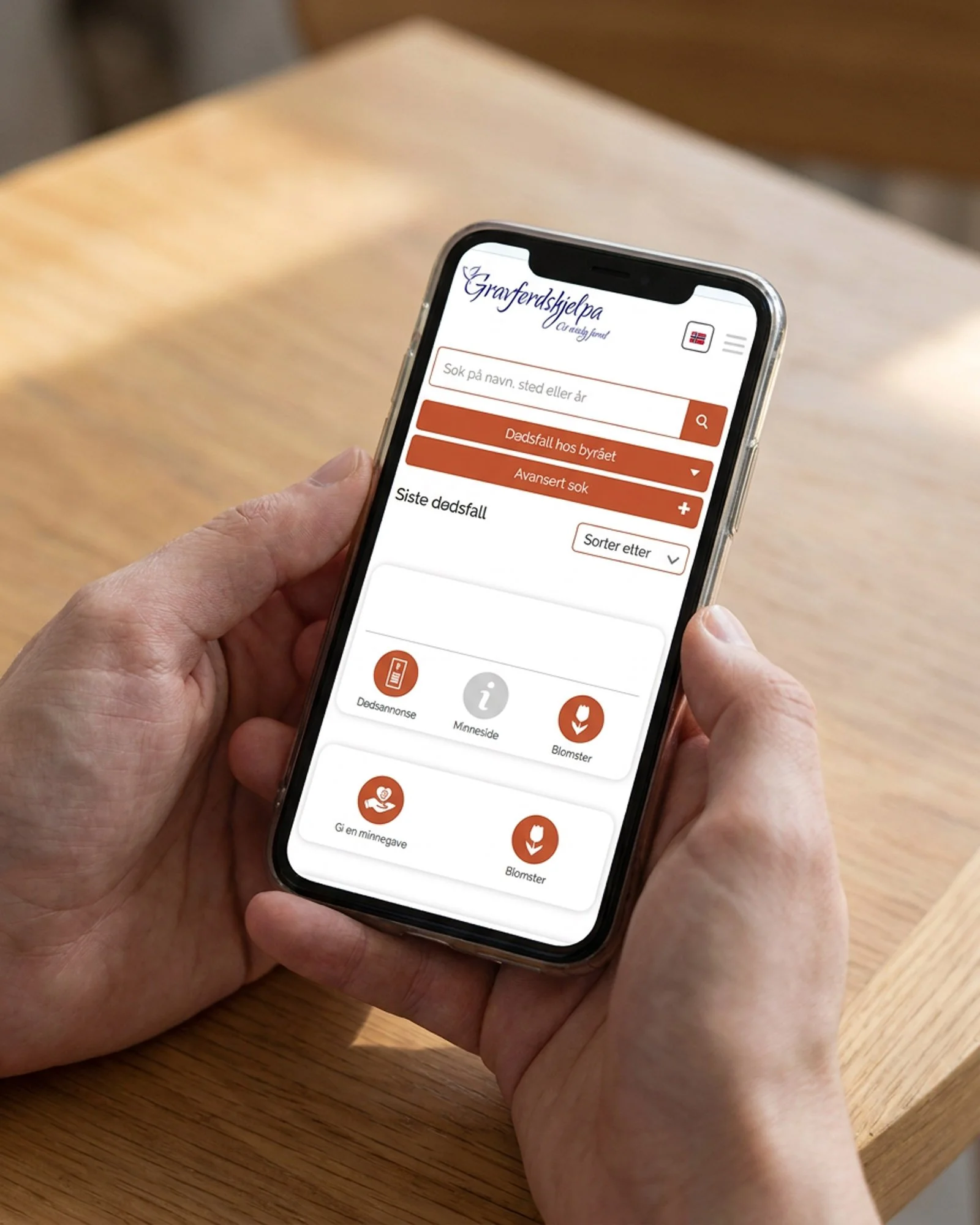 En person holder en smarttelefon med en applikasjon for å finne hjelp i en krisesituasjon, med ulike funksjoner som dødsannonse, minneside og blomster.