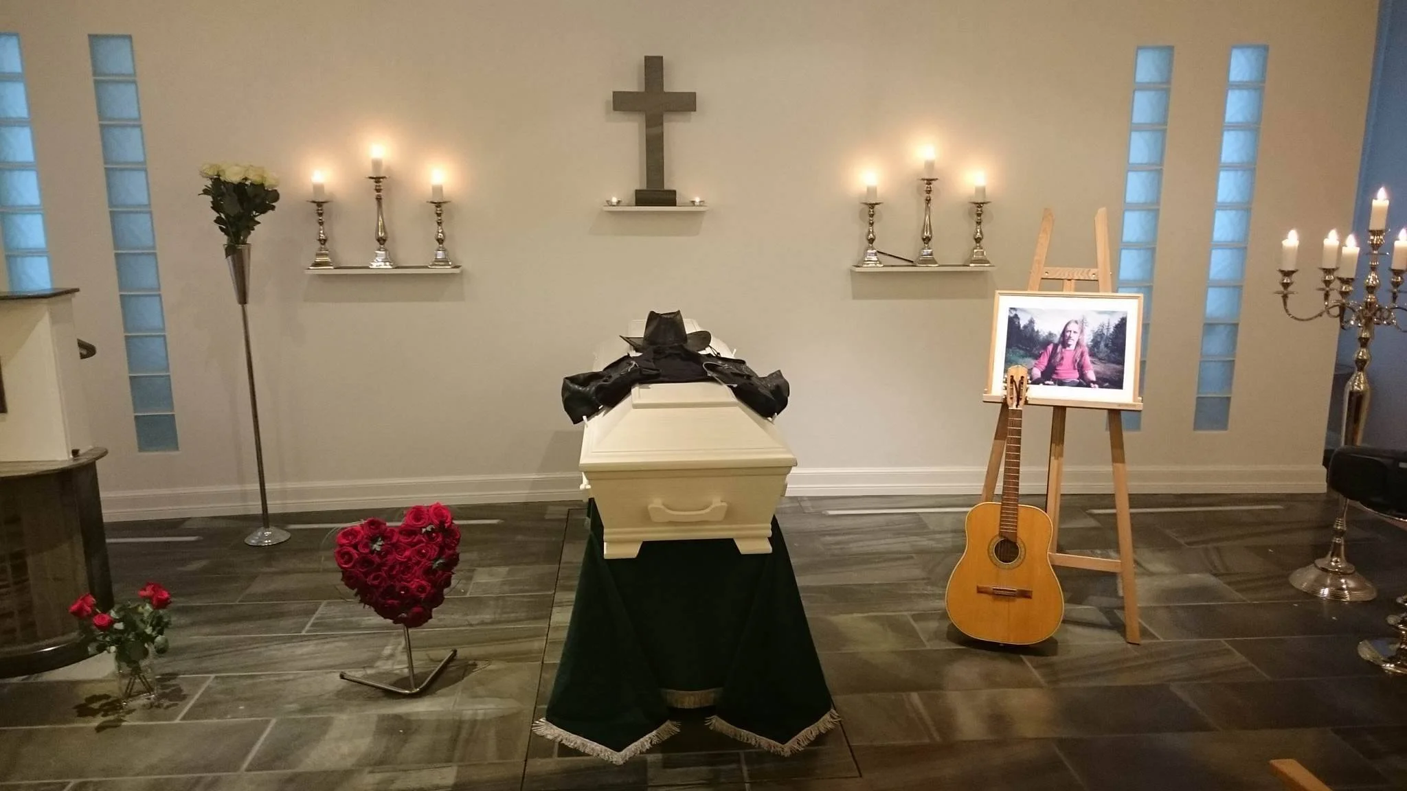 Et dødsleie med åpen kiste, blomster, tente lys, et kors på veggen, en gitar, og et bilde av en person i en utendørs setting.