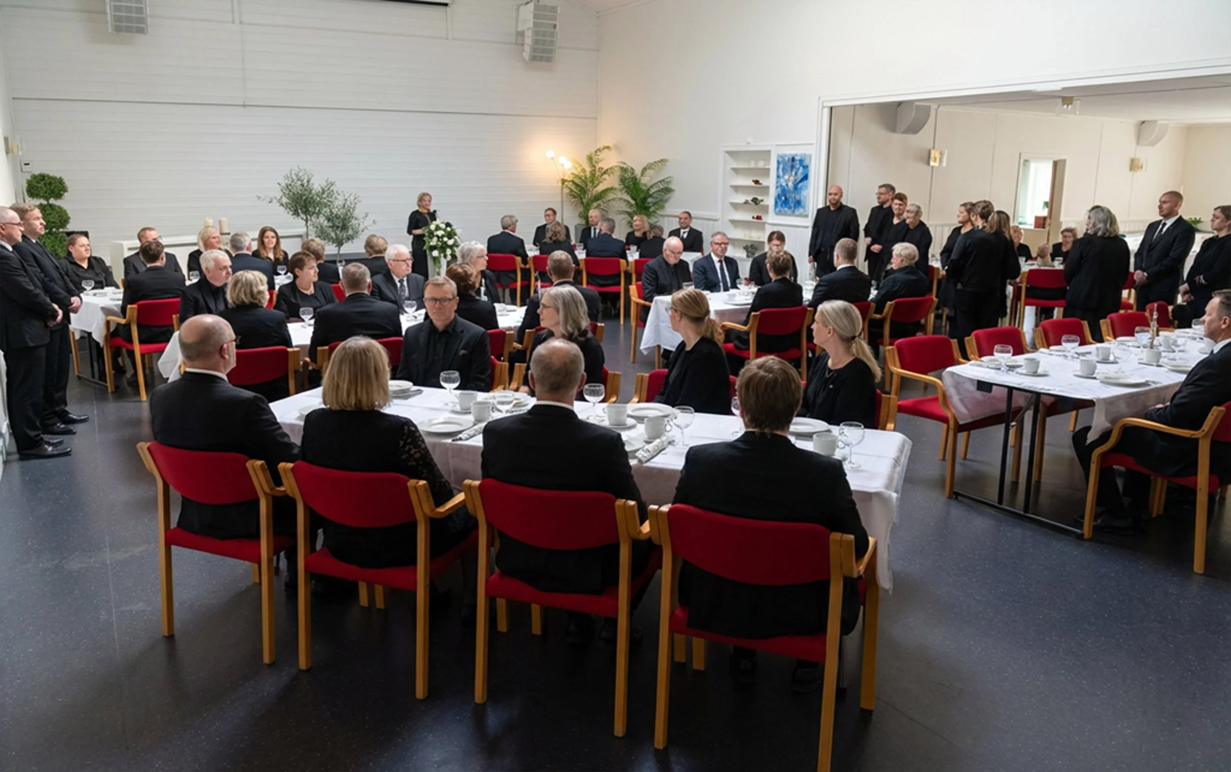 En gruppe mennesker i formelle klær samles i et rom med hvite vegger, rundt bord dekket med servise og glass. Noen står, mens andre sitter og lytter til en kvinne som snakker.