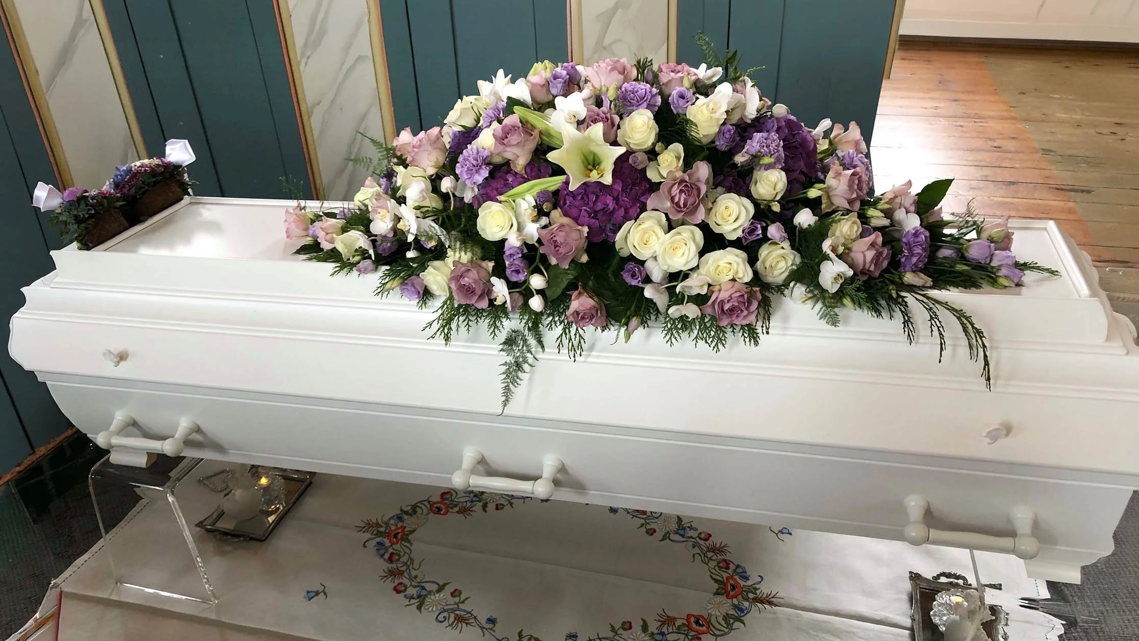 Kista dekket med en blomsterarrangement av hvite, lilla og rosa blomster, plassert på en hvit koffert i et rom med trepanel og en treparkett