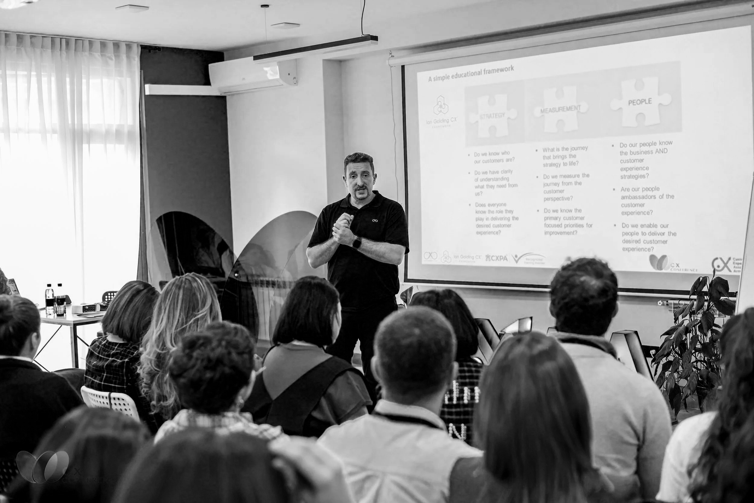 A man in a black shirt giving a presentation to an audience in a conference room. There is a large screen with a slide titled 'A simple educational framework' showing puzzle pieces labeled 'Strategy,' 'Measurement,' and 'People.' The audience is seated and paying attention.