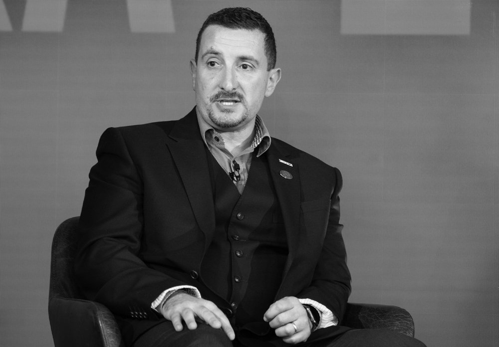 Man in a suit sitting in a chair, speaking in an interview or panel discussion, in black and white.