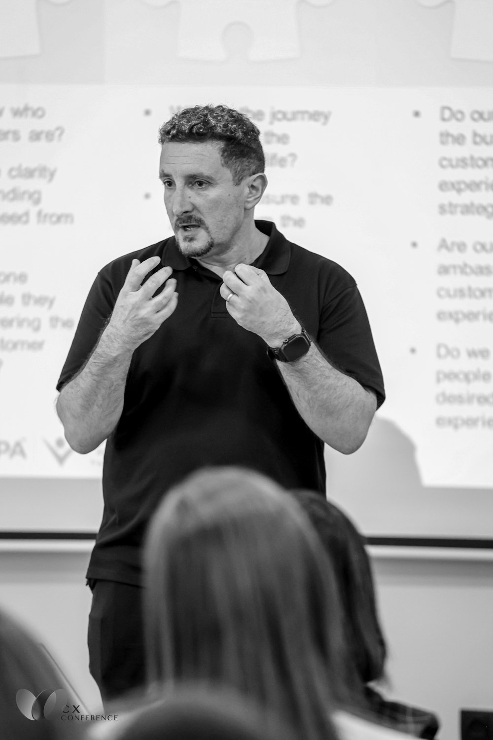 A man, wearing a black polo shirt, is speaking and gesturing with his hands in a classroom or conference room setting. There is a presentation slide with text behind him and audience members in the foreground.