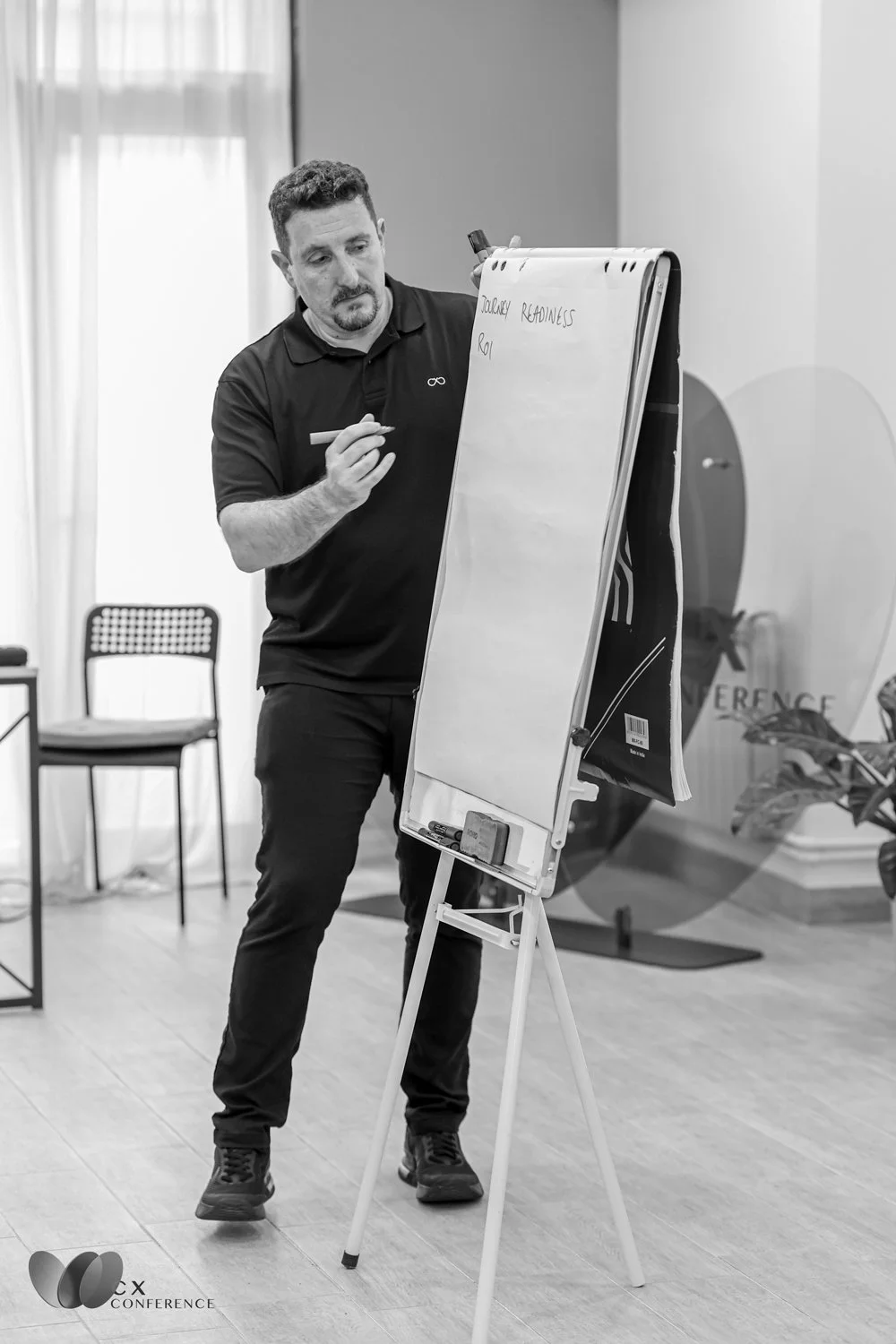 Man standing in front of a whiteboard during a conference, writing notes with a marker. The room has chairs and a plant, with a logo on the bottom left corner that reads 'CX Conference'.