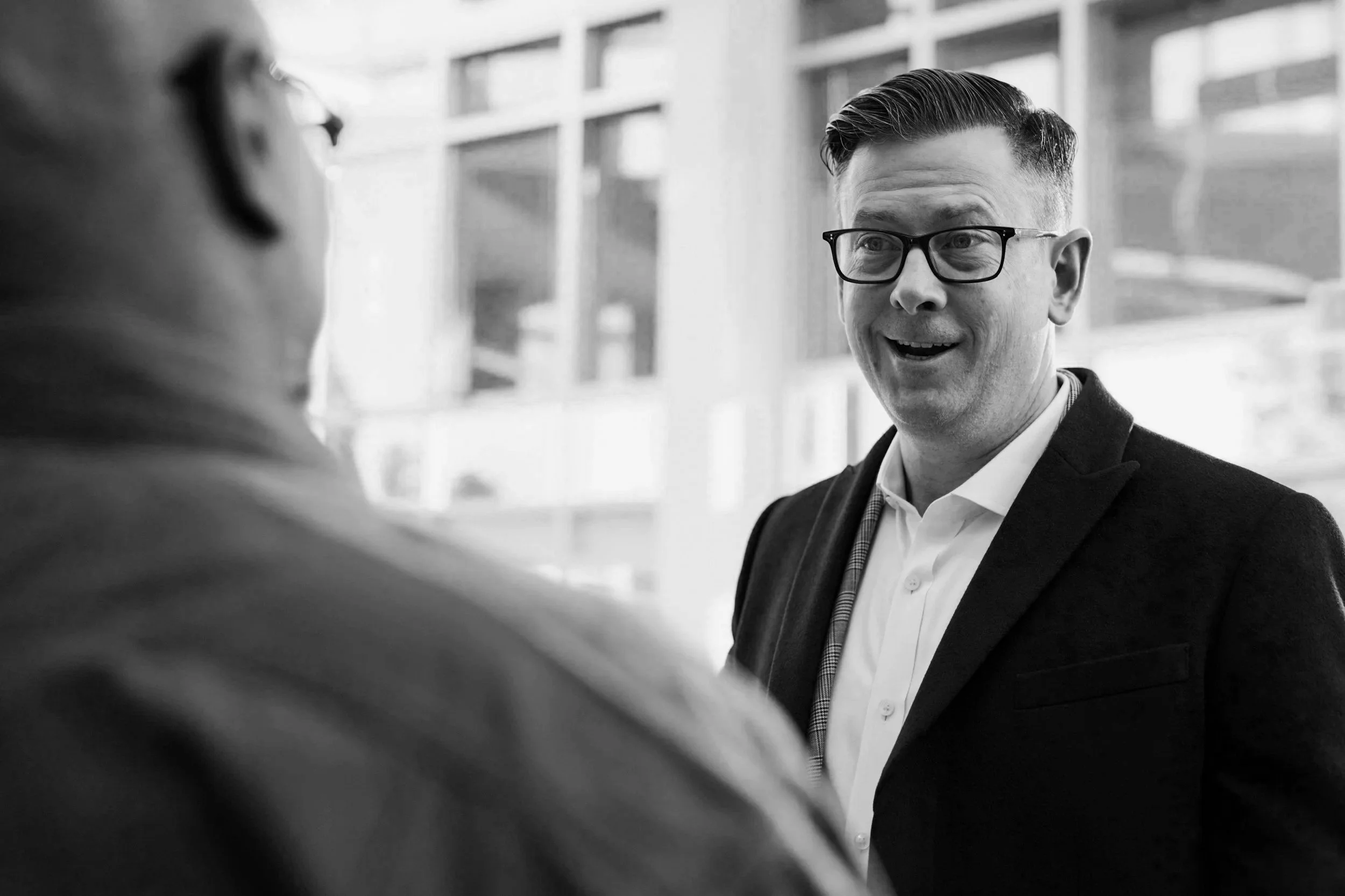 A man wearing glasses and a suit is talking to a woman with glasses in an office setting, with large windows in the background.