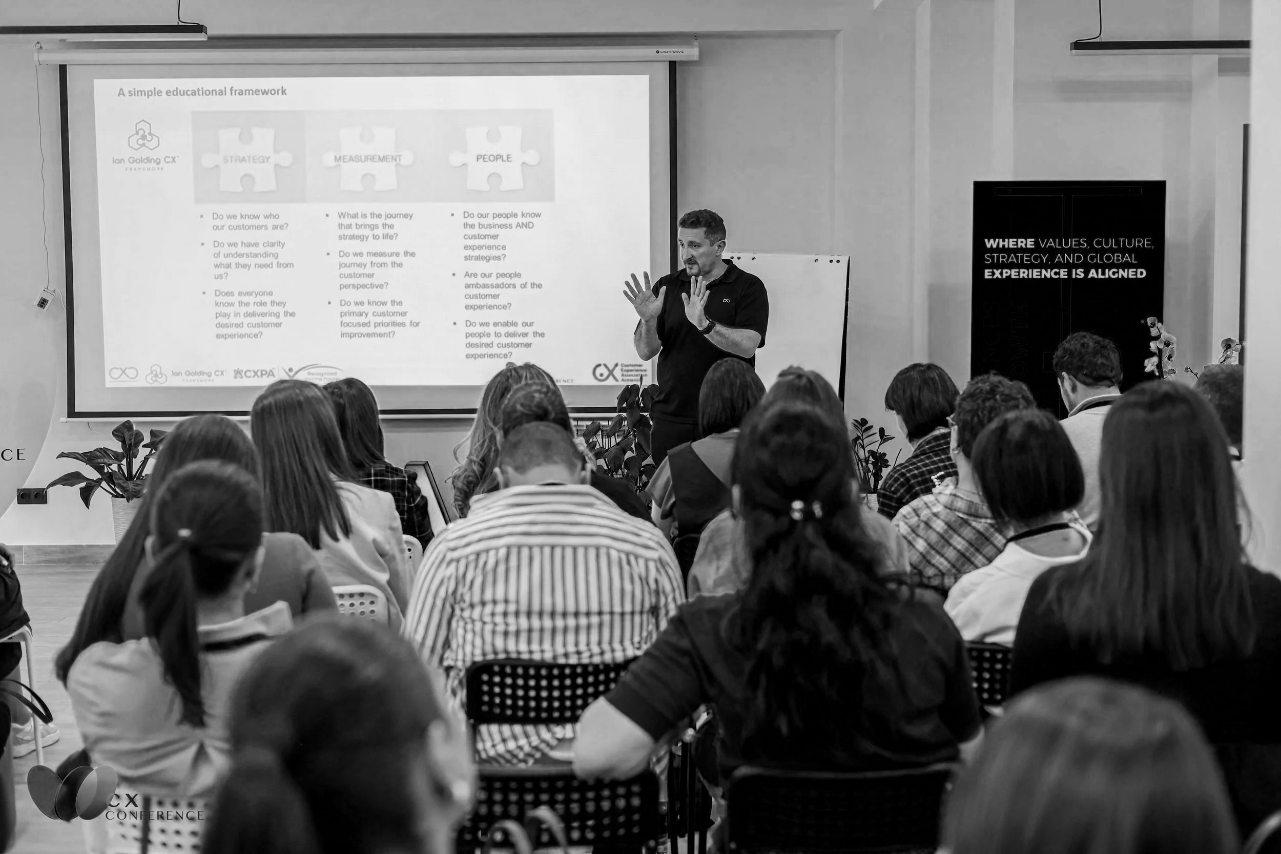 A man presenting at a conference in front of a large screen with presentation slides, with an audience seated and listening.