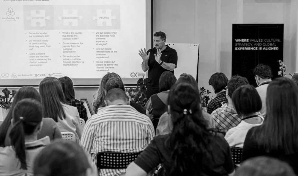 Man giving a presentation to a group of people in a classroom or conference room with a projected slide and a sign on the wall that reads, 'Where values, culture, strategy, and global experience is aligned.'