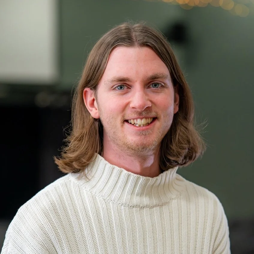 A smiling man with long, wavy brown hair and blue eyes, wearing a white turtleneck sweater, indoors with a blurred background.