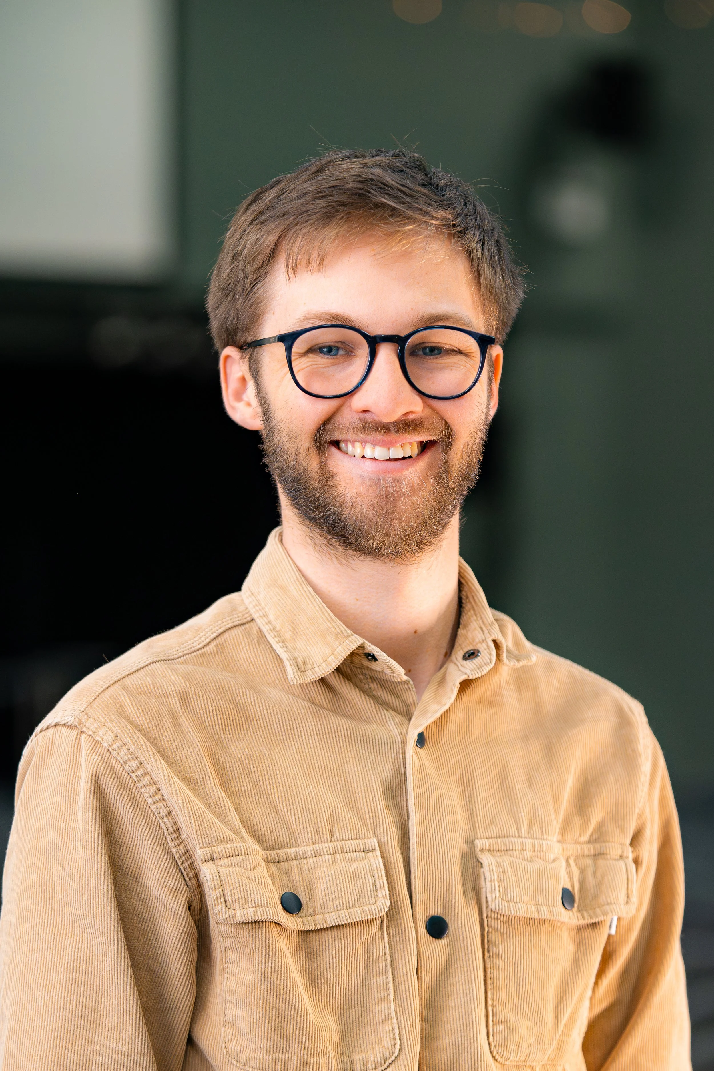 A young man with light brown hair, glasses, and a beard smiling, wearing a tan button-up shirt, standing in front of a blurred, dark background.