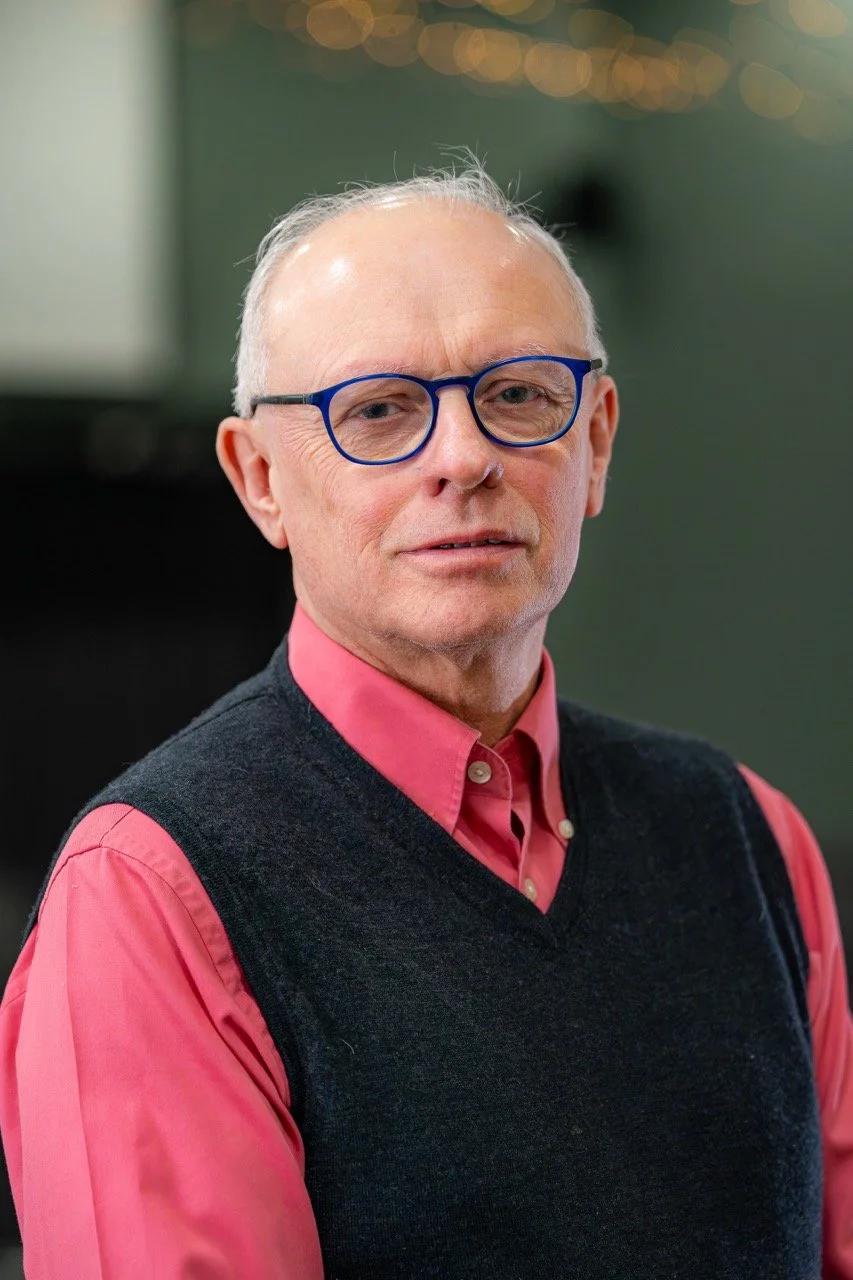 A man with gray hair, wearing blue glasses, a pink shirt, and a black sweater vest, looking at the camera with a slight smile. The background is blurred with circular lights.