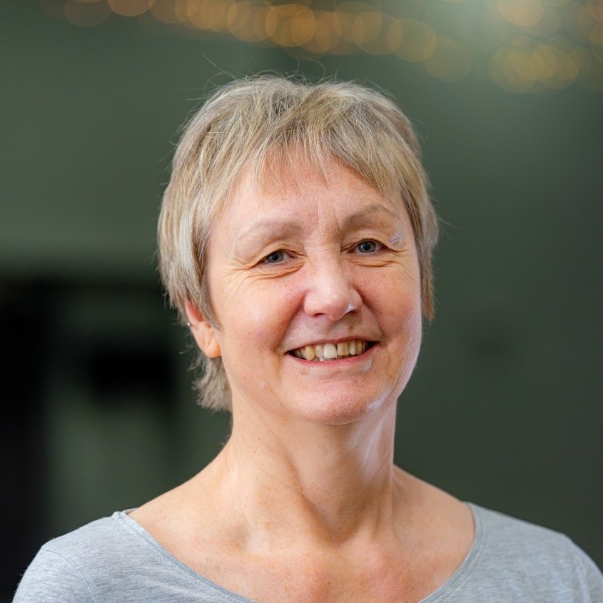 A smiling middle-aged woman with short blonde hair, wearing a light grey top, standing indoors with blurred background.