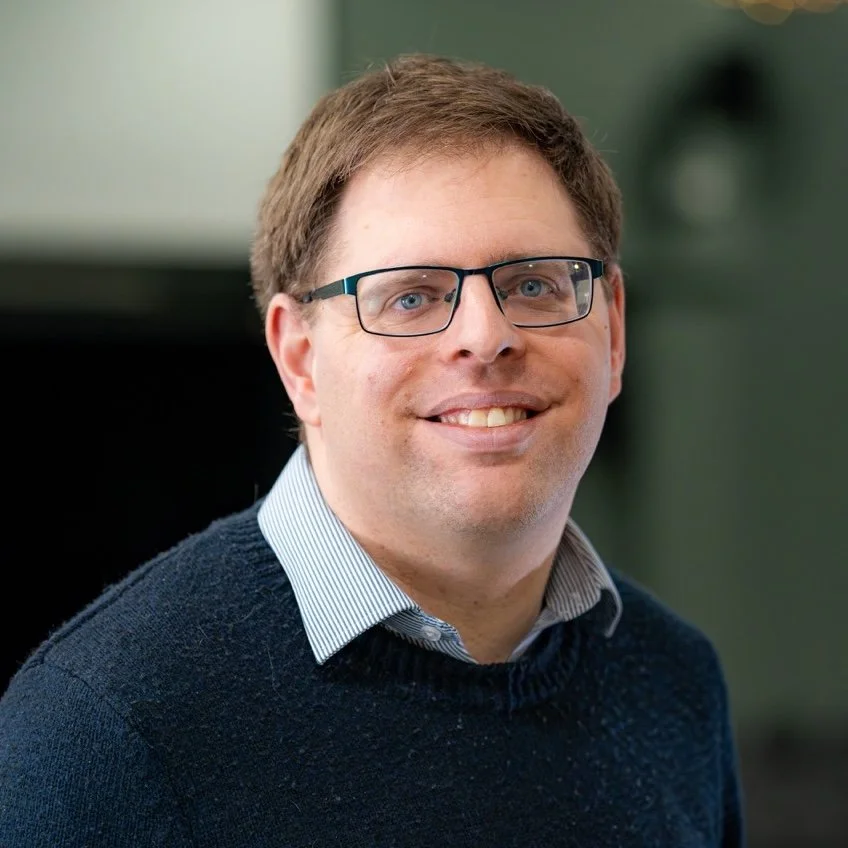 A man with brown hair, glasses, and a light blue collared shirt smiling in an indoor setting.